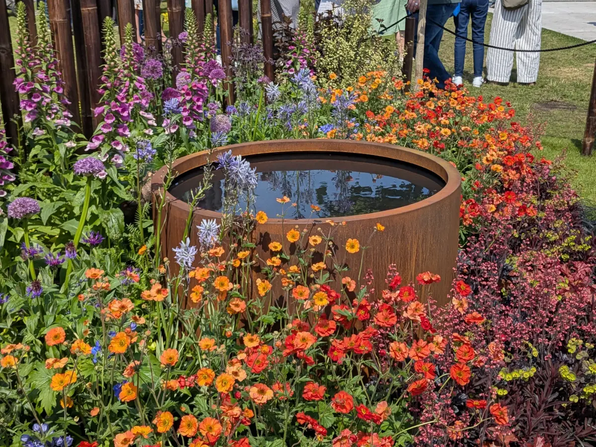 A vibrant garden border features a round, rusty corten steel water feature, surrounded by tall purple, blue, orange, and red flowers.