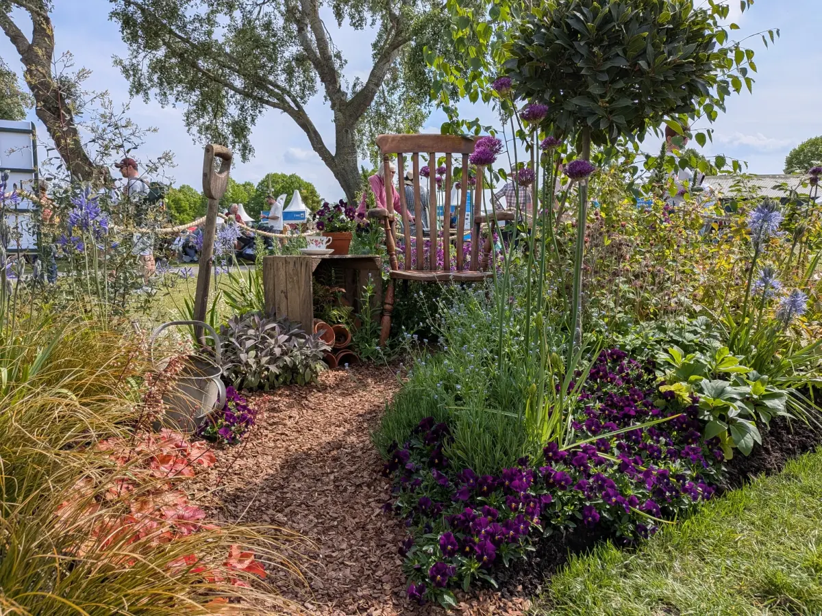 A rustic garden border features a wooden chair and plinth with a teacup, surrounded by a woodchip path and abundant purple, blue, and coral-toned planting.