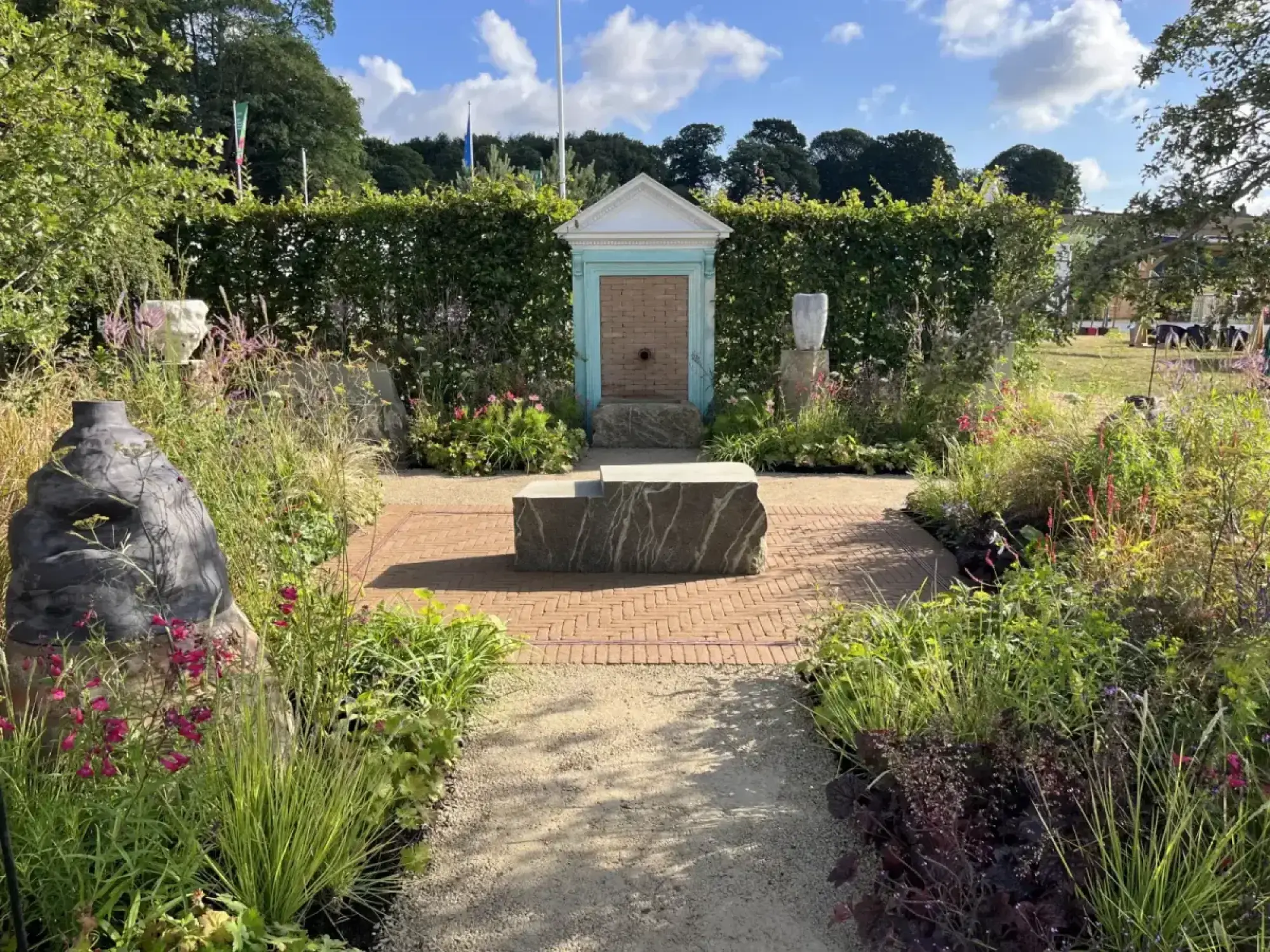 A Young Designer Garden presents a pale blue classical fountain, central veined stone block, and sculptures in naturalistic pink-flowered planting.
