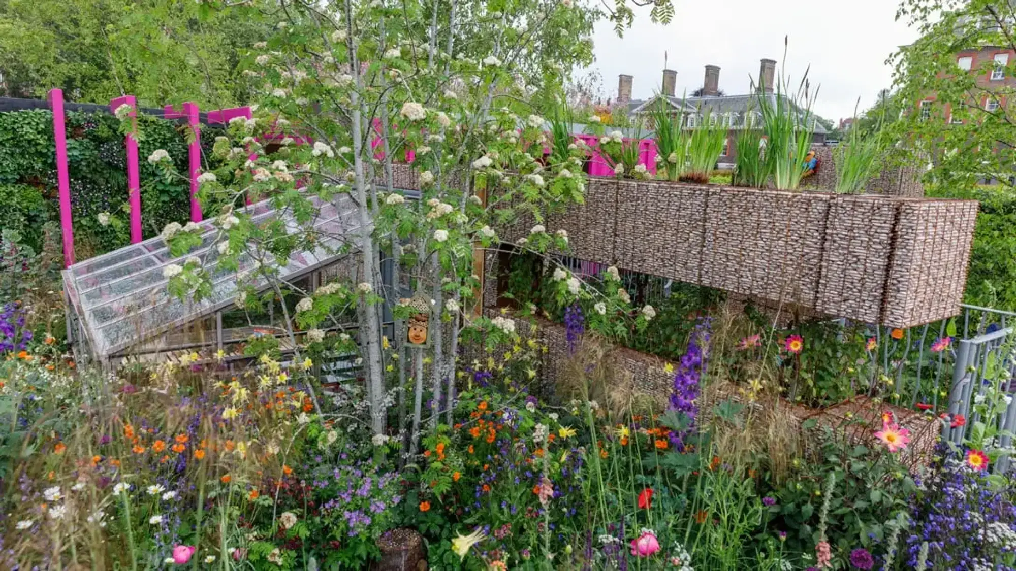 A colourful children's garden features a tilted greenhouse, vibrant pink structural posts, multi-level woven planters, and diverse flowering plants.