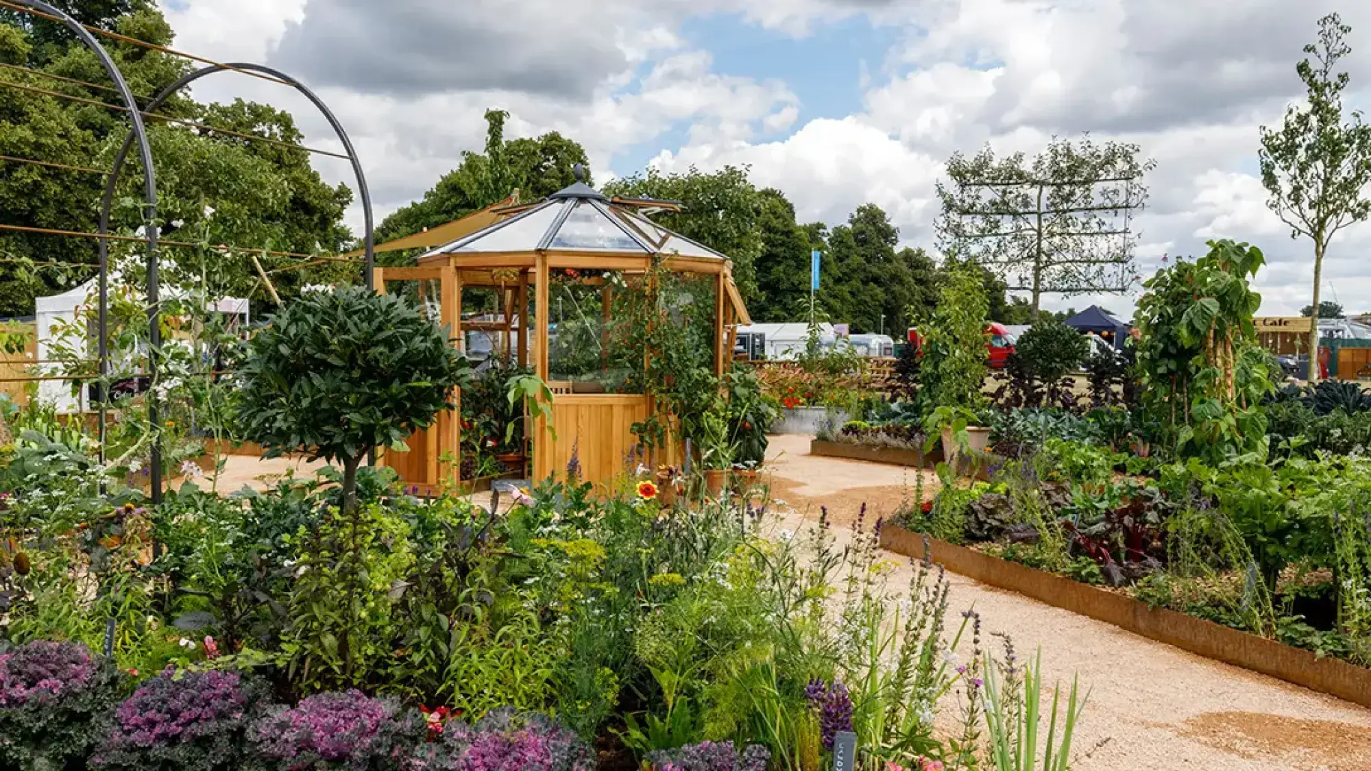 A vibrant market garden features an octagonal wooden greenhouse, gravel paths, corten steel raised beds, and abundant edible plants including purple kale and climbing beans.