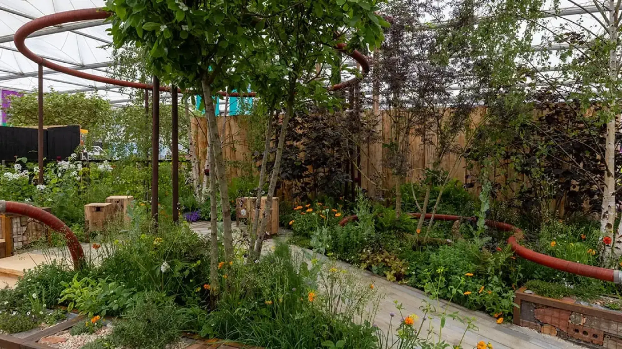 A naturalistic garden features a prominent curved red pipe structure winding through mixed planting, with wooden log seats and a wood plank pathway.