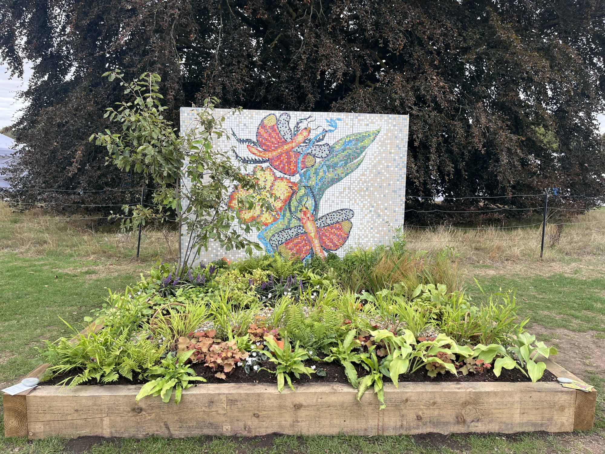 A beautiful borders garden featuring a large white mosaic panel with colourful butterflies and lush green and purple planting in a timber raised bed.