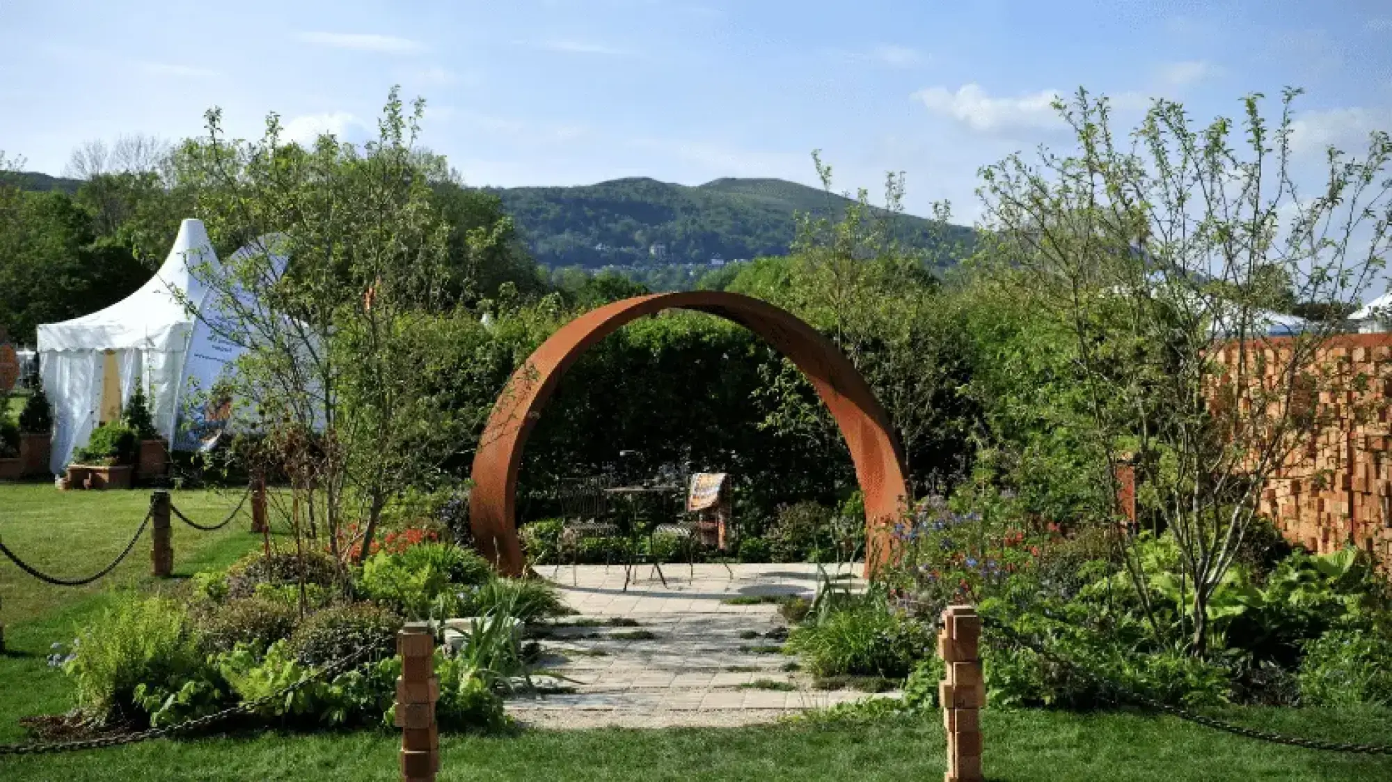 A contemporary show garden features a large rusted arch, a flagstone path, outdoor seating, lush mixed planting, and a wall of recycled bricks.