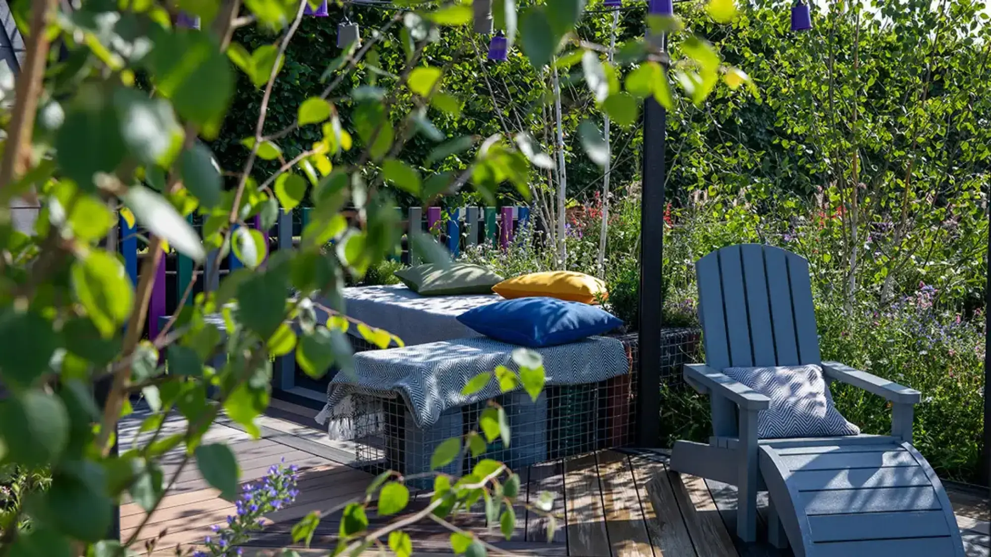 A grey Adirondack chair and a gabion bench of colourful recycled plastic on decking, surrounded by lush planting and a vibrant fence.