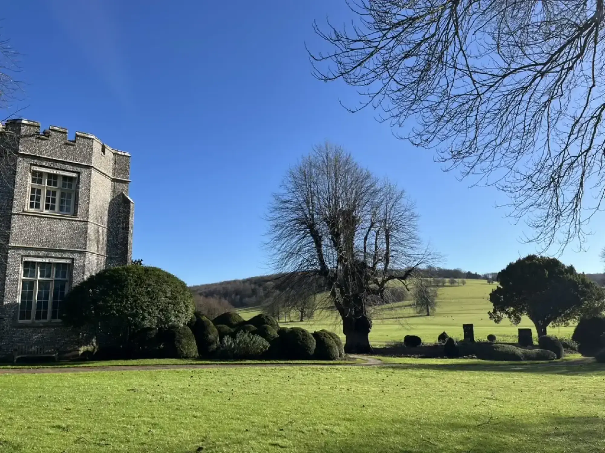 West Dean Gardens showcases a grand flint stone mansion with battlements, set amidst an expansive green lawn, sculpted topiary, and large mature trees.