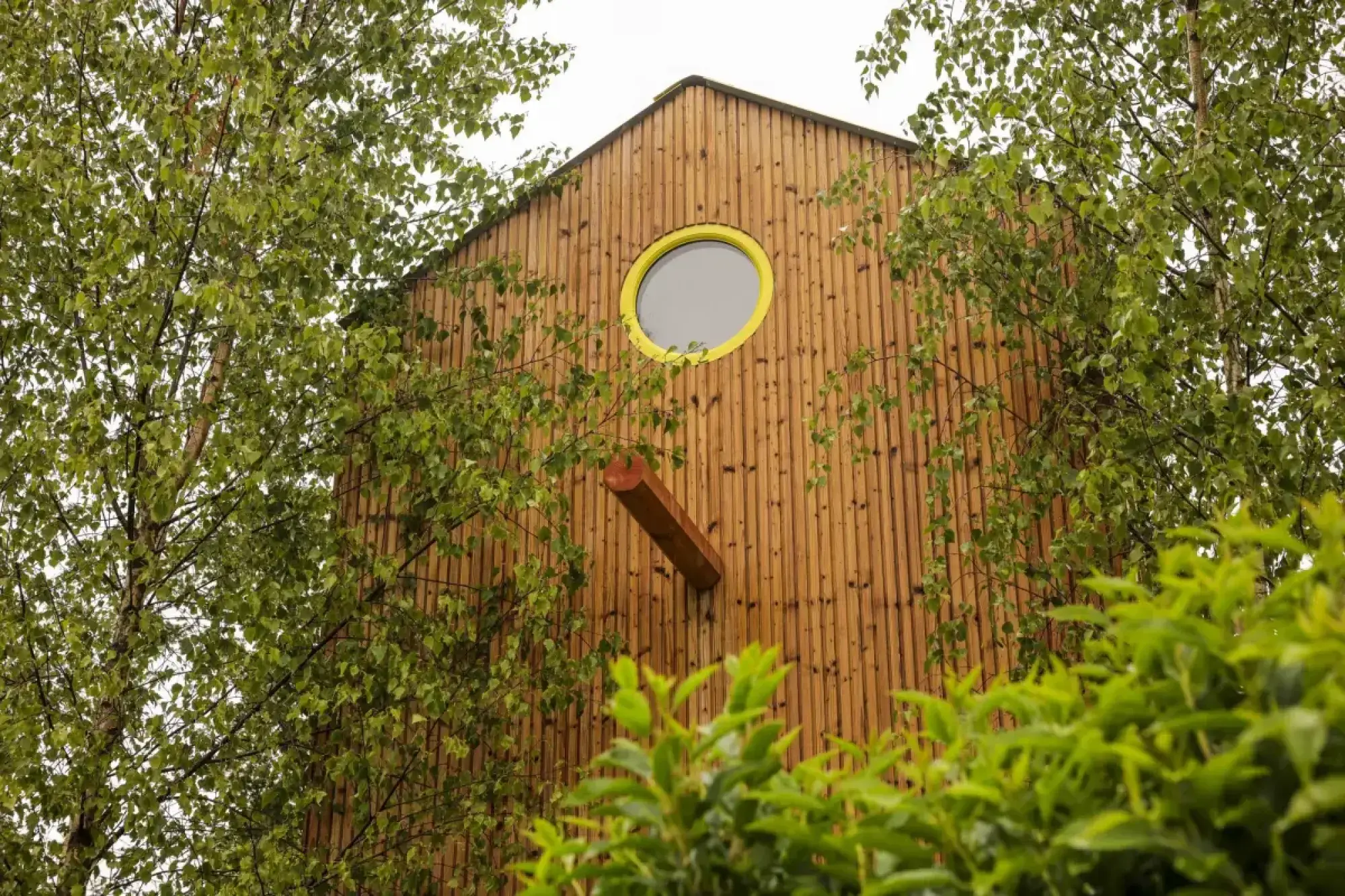 A tall, narrow wooden structure with vertical cladding features a yellow-framed round window and a protruding wooden beam, surrounded by green trees and shrubs.