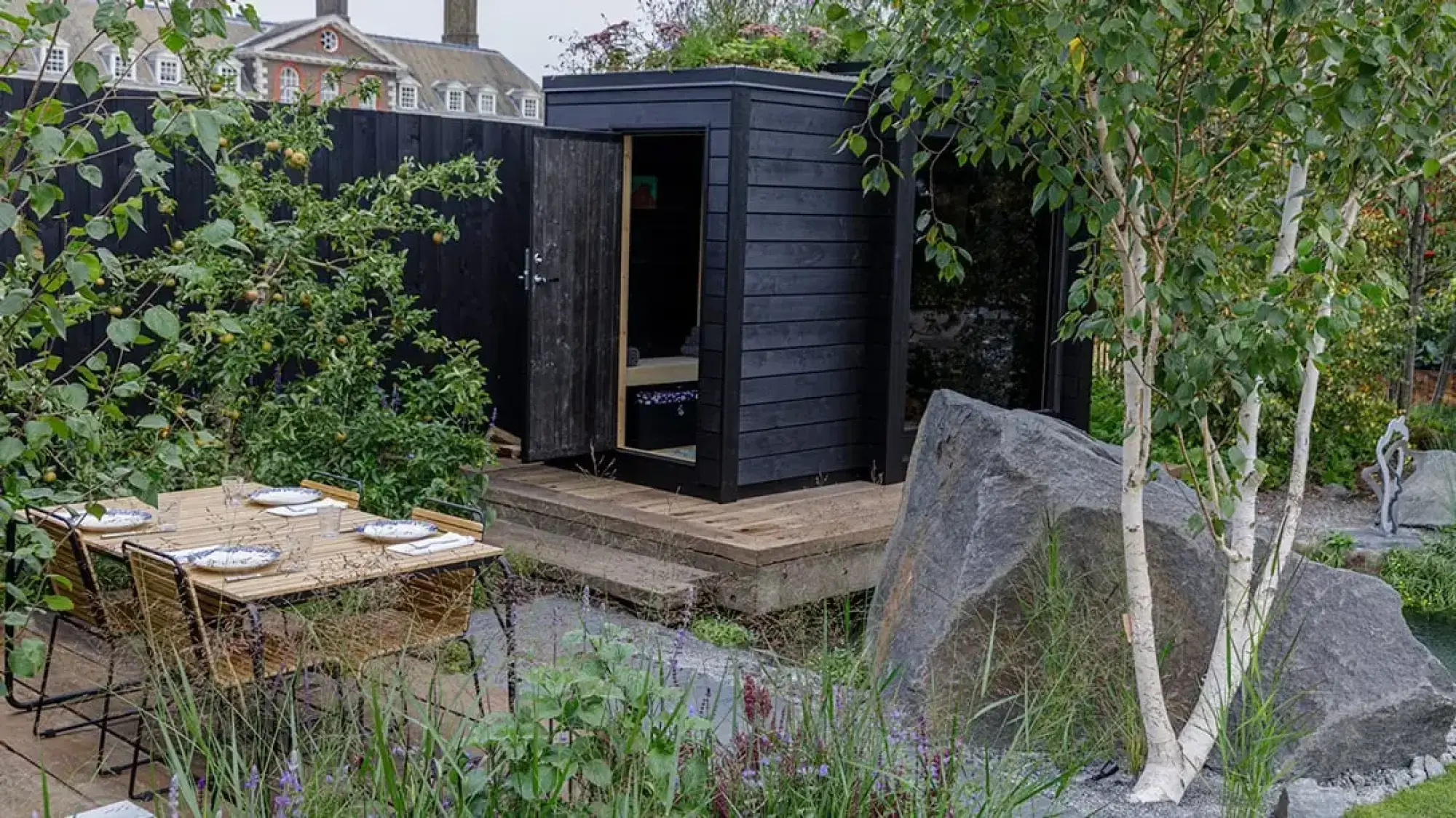 A tranquil Nordic heritage garden featuring a black timber sauna cabin with a green roof, set among large granite boulders and multi-stemmed birch trees, with a naturalistic pond and an outdoor dining area.