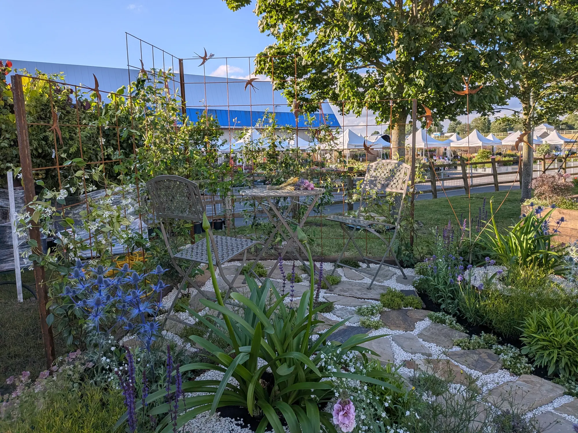 A beautiful border garden with a rustic metal trellis, bird sculptures, a bistro set, a stone path, and blue, purple, and white flowering plants.