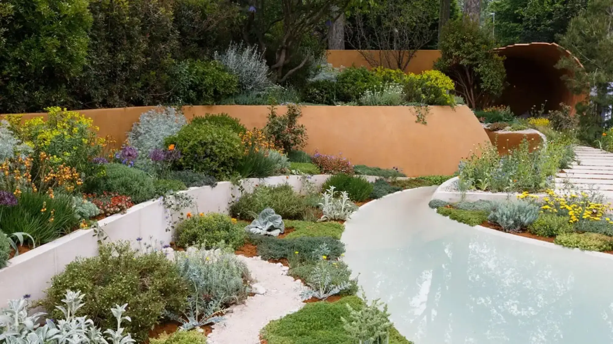 The Dubai Majlis Garden showcases a curved orange wall, a flowing white water channel, and a rusted metal tunnel, amidst colourful drought-tolerant planting.
