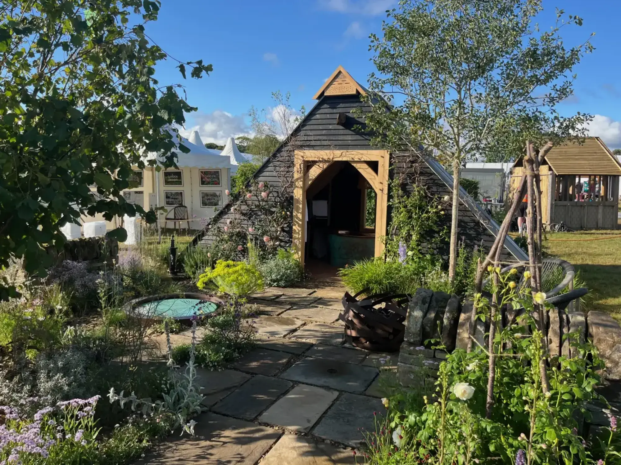 A rustic show garden with a pyramid-shaped, black-shingled barn, a flagstone path, a circular pond, a dry stone wall, and naturalistic planting.