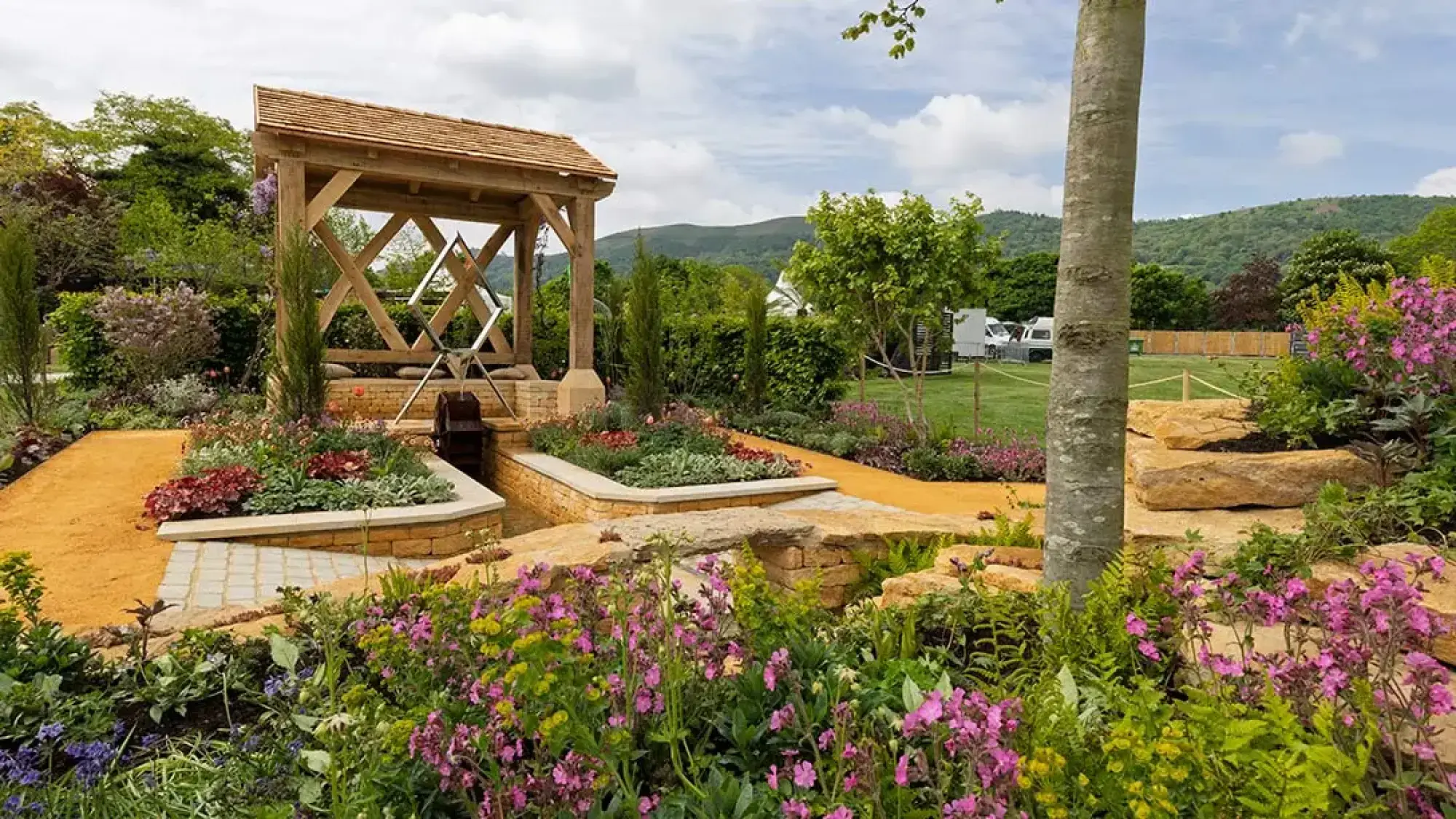 A show garden features a timber-framed watermill structure with a diamond sculpture, tiered Cotswold stone walls, and a golden gravel path surrounded by lush pink and purple flowering plants.