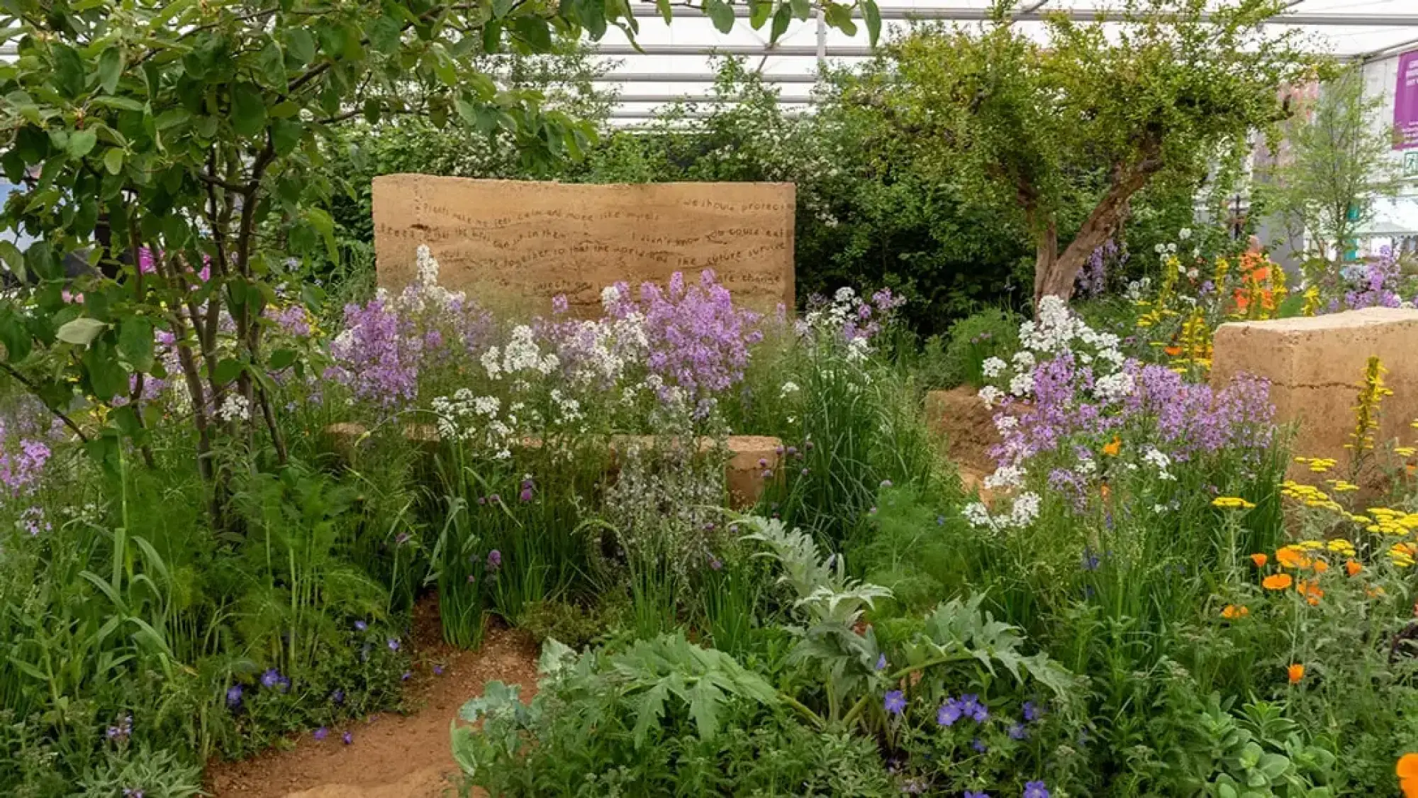 A naturalistic garden features a rammed earth wall with etched text, surrounded by vibrant purple, white, yellow, and orange wildflowers and diverse green plants.