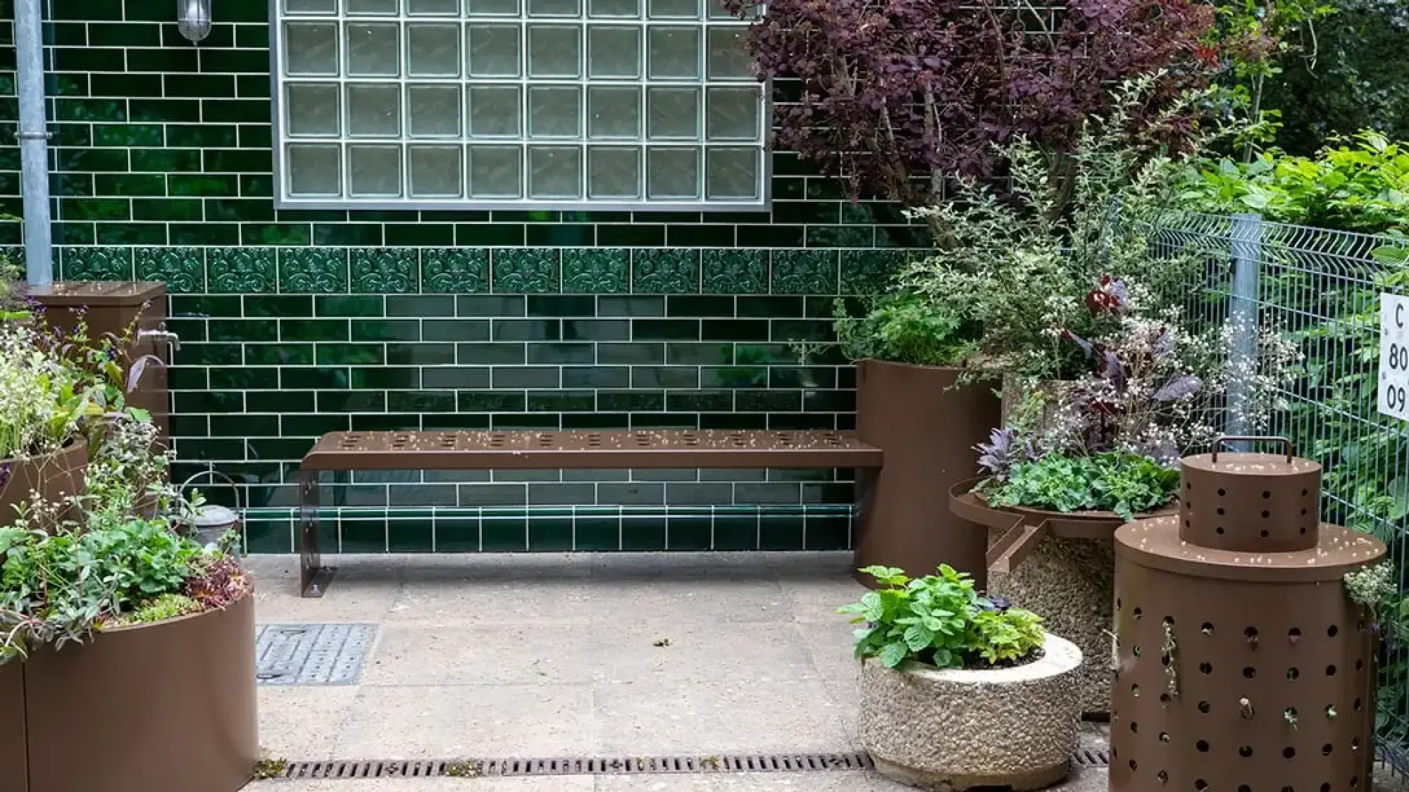 A container garden with a dark green tiled wall, glass block window, brown metal bench, and planters with mixed green and purple plants, plus a rain barrel.