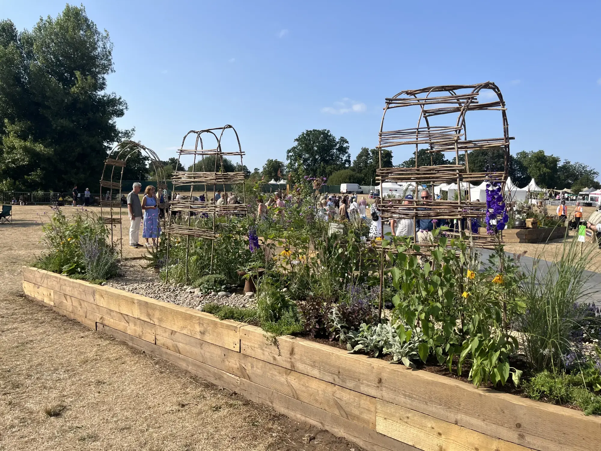 A long border garden featuring two large, open willow arches, a raised timber planter, and a mix of green, purple, and yellow herbaceous plants with ornamental grasses and gravel.
