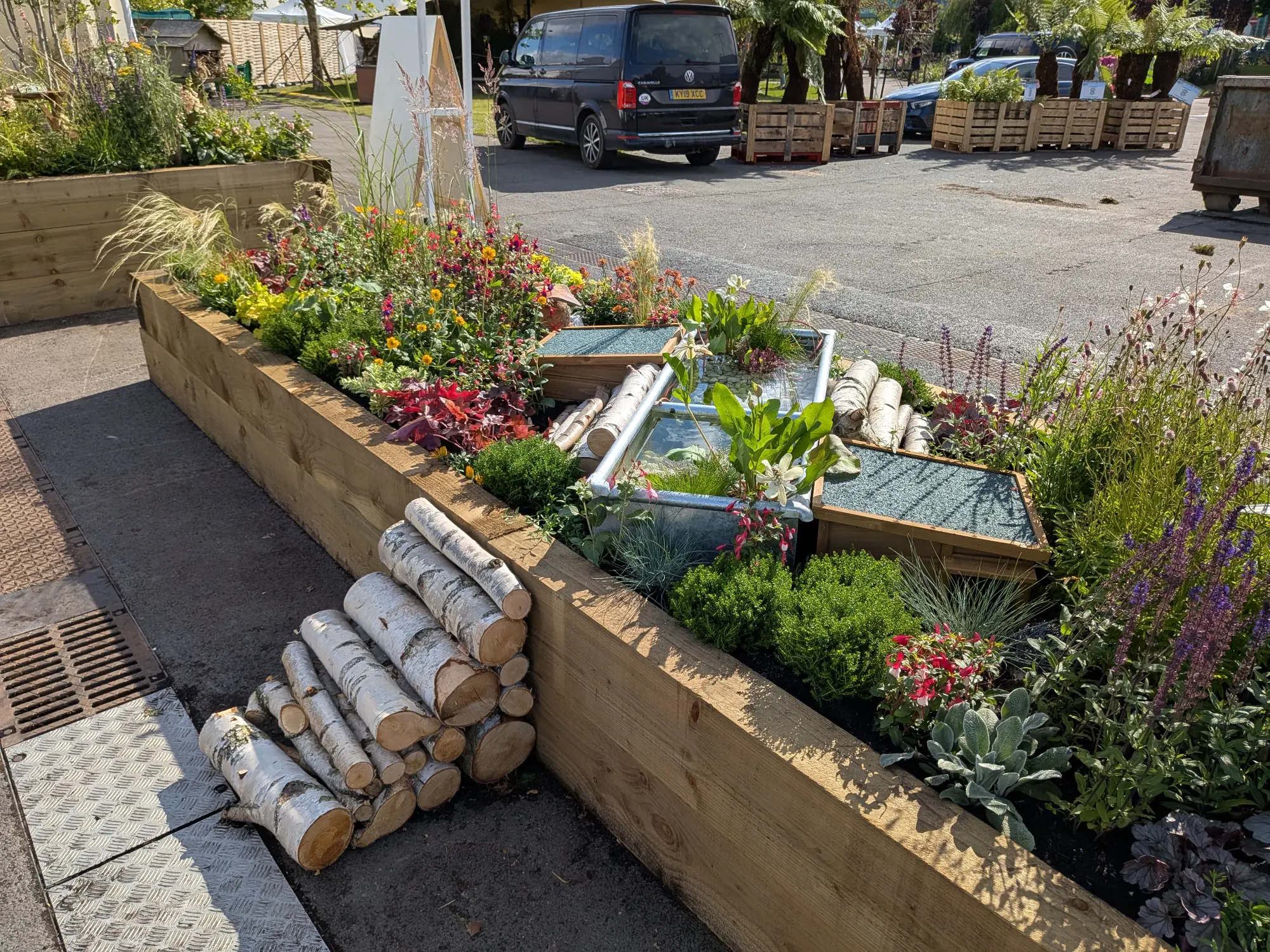 A long timber raised border garden featuring two rectangular water troughs, one with a wooden cover, surrounded by mixed colourful planting and stacked birch logs.