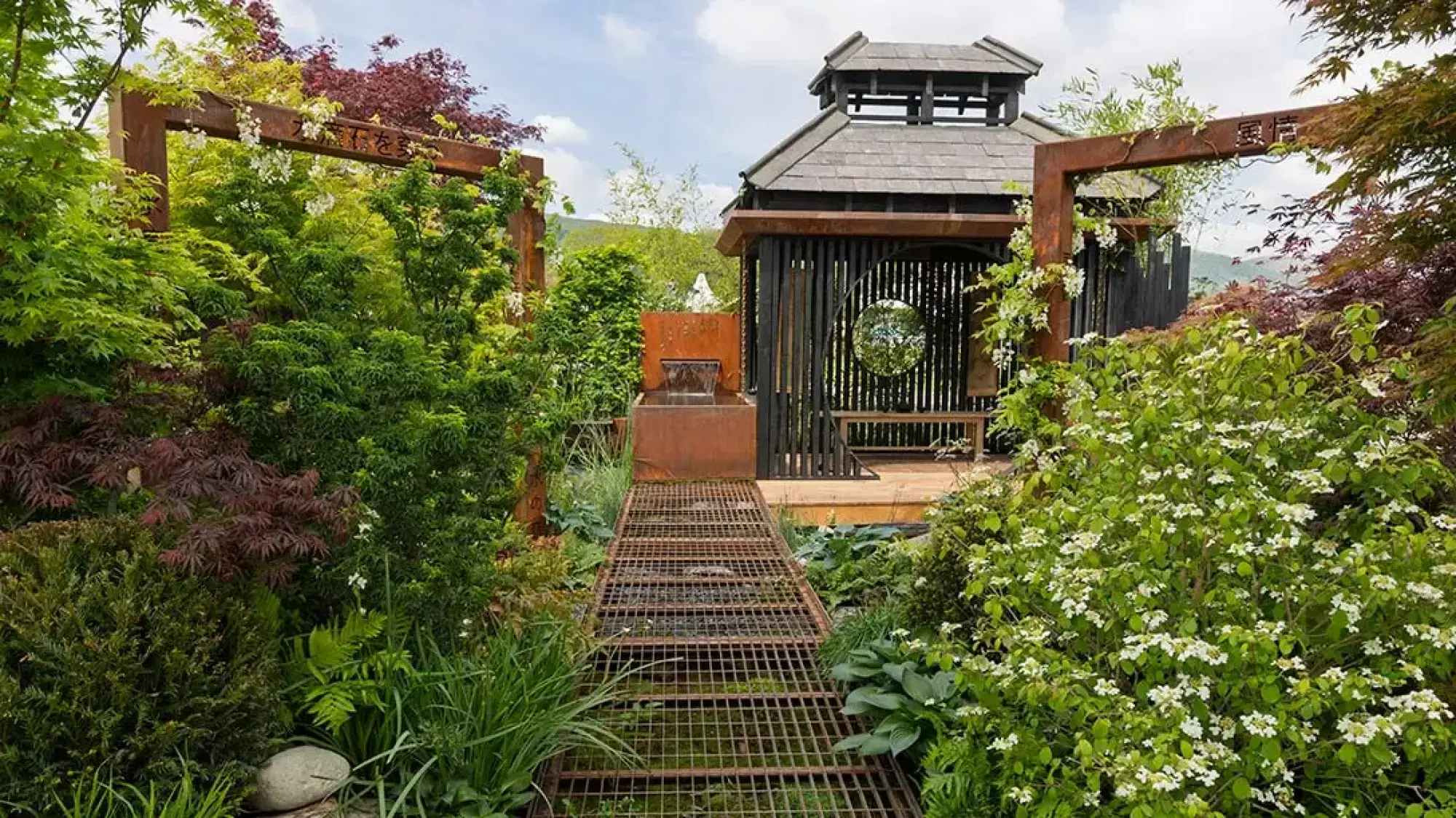A rain garden with a dark wood pavilion, corten steel archway and waterfall, metal grid path over water, and lush green plants.