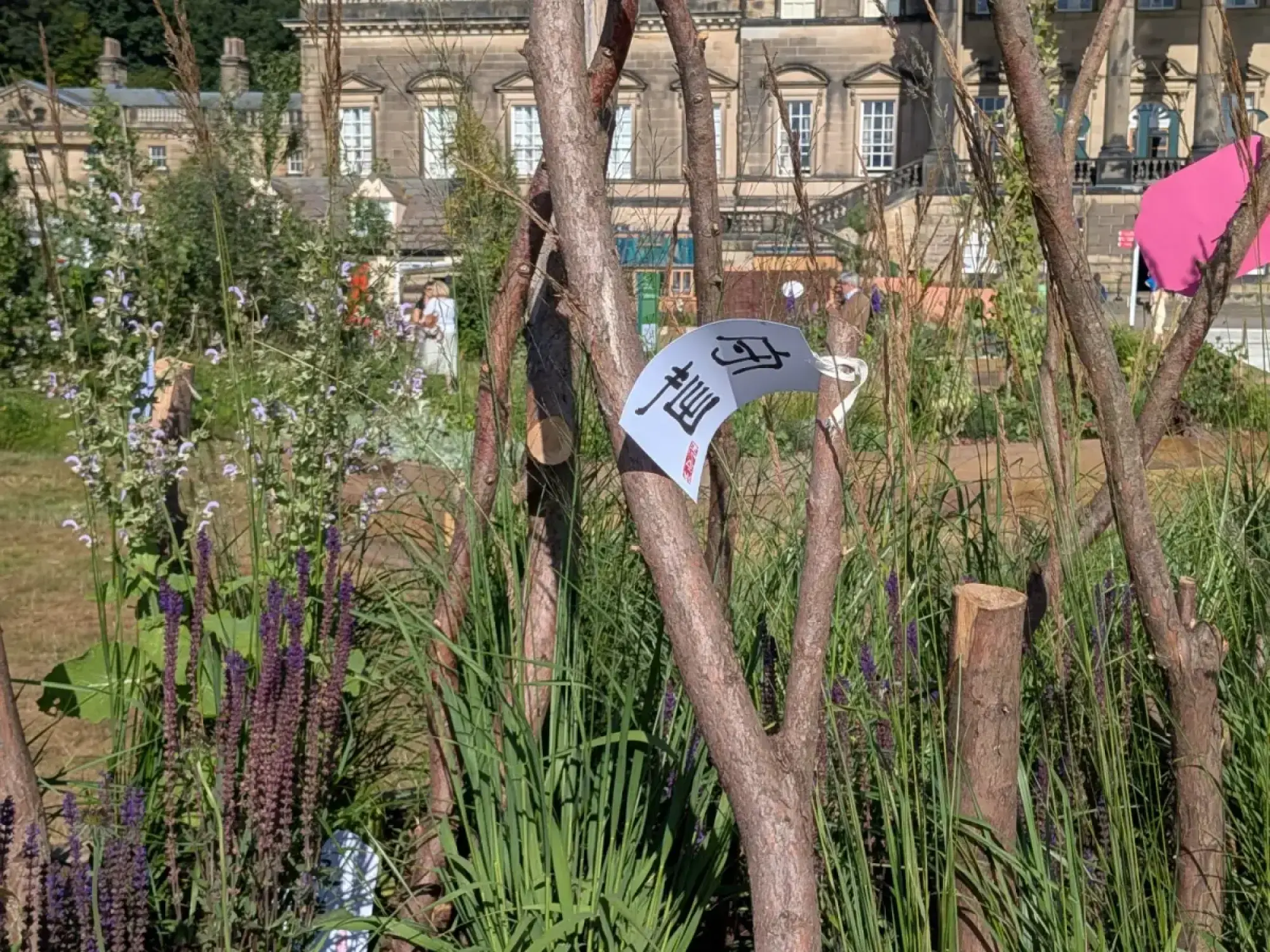 A naturalistic long border garden featuring tall grasses, spiky purple and white flowers, cut wood branches, and a white fan ornament with calligraphy.