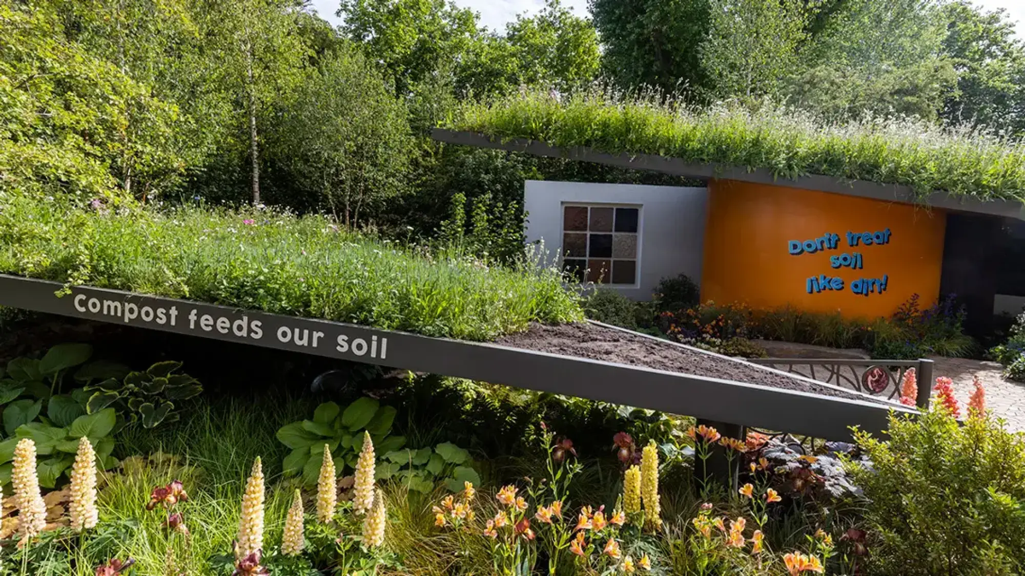 A modern educational garden displays green roofs with environmental messages, an orange wall, a window showing soil types, and vibrant mixed planting.