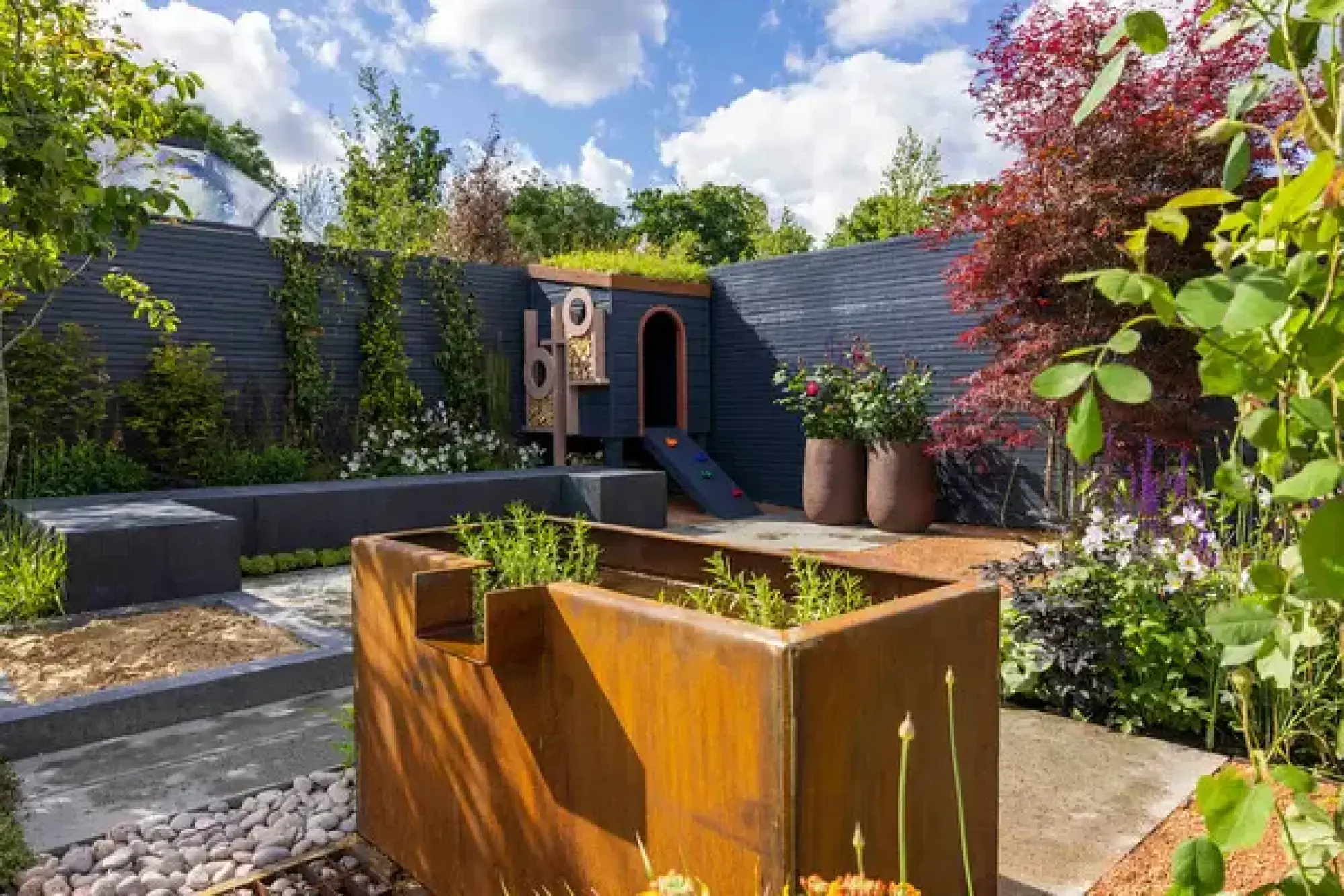 A contemporary family garden with a dark blue playhouse, green roof, climbing ramp, sandpit, and a corten steel planter among diverse planting.