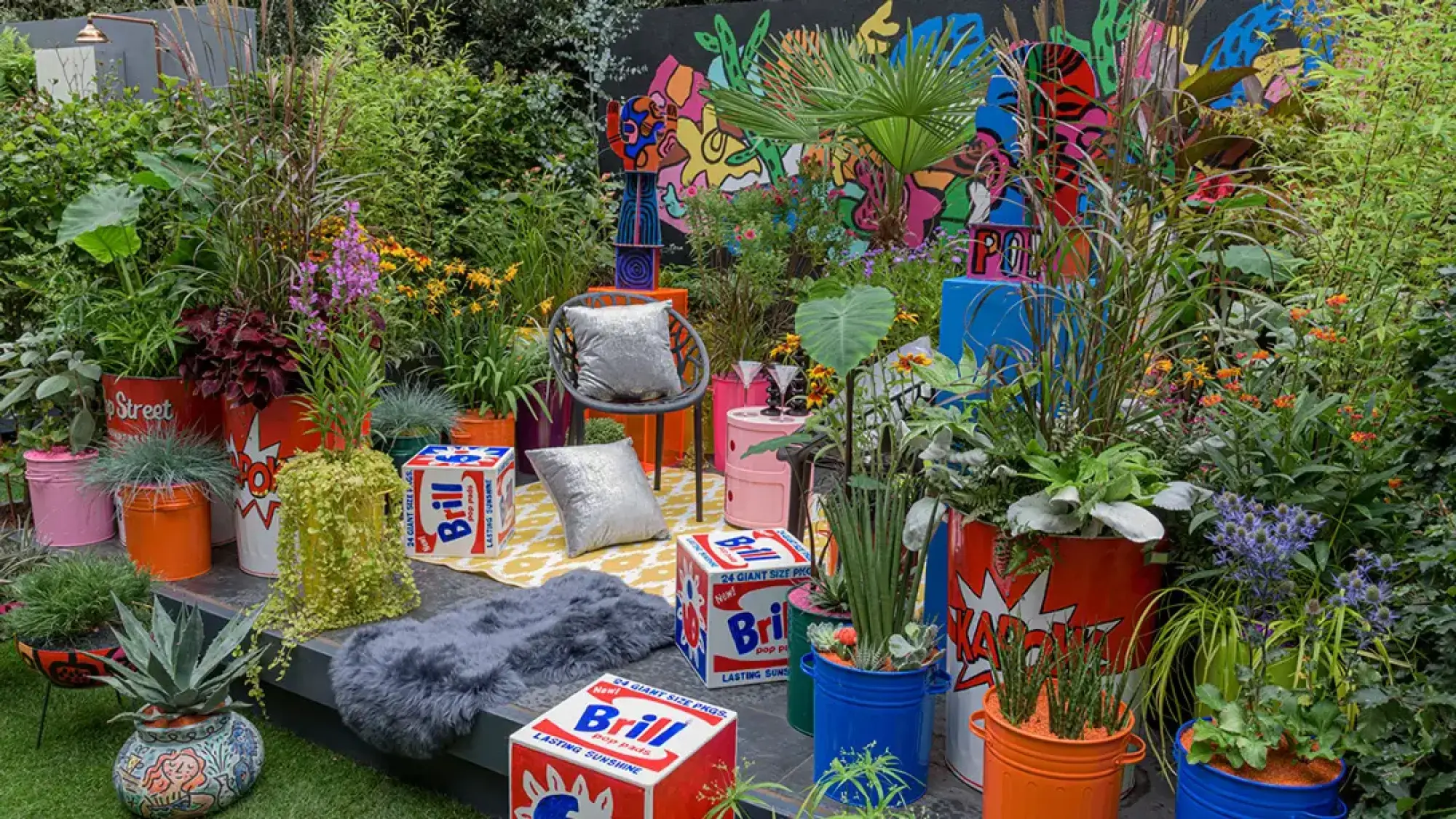 A vibrant pop art container garden featuring brightly painted comic-style barrels, Brill boxes, a colourful mural, modern seating, and lush planting.