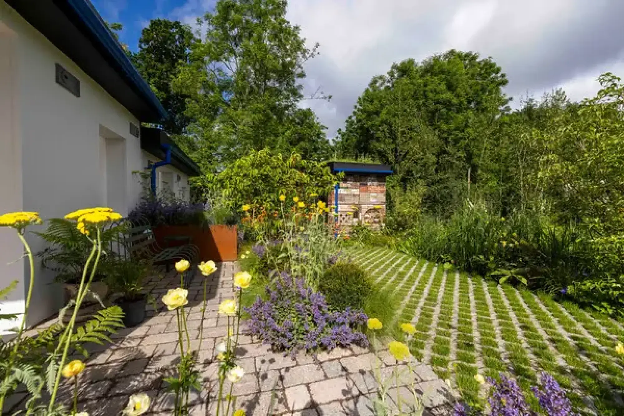 A show garden features a large insect hotel, a white building, brick paver path, a grass grid, and bright yellow and purple flowers.