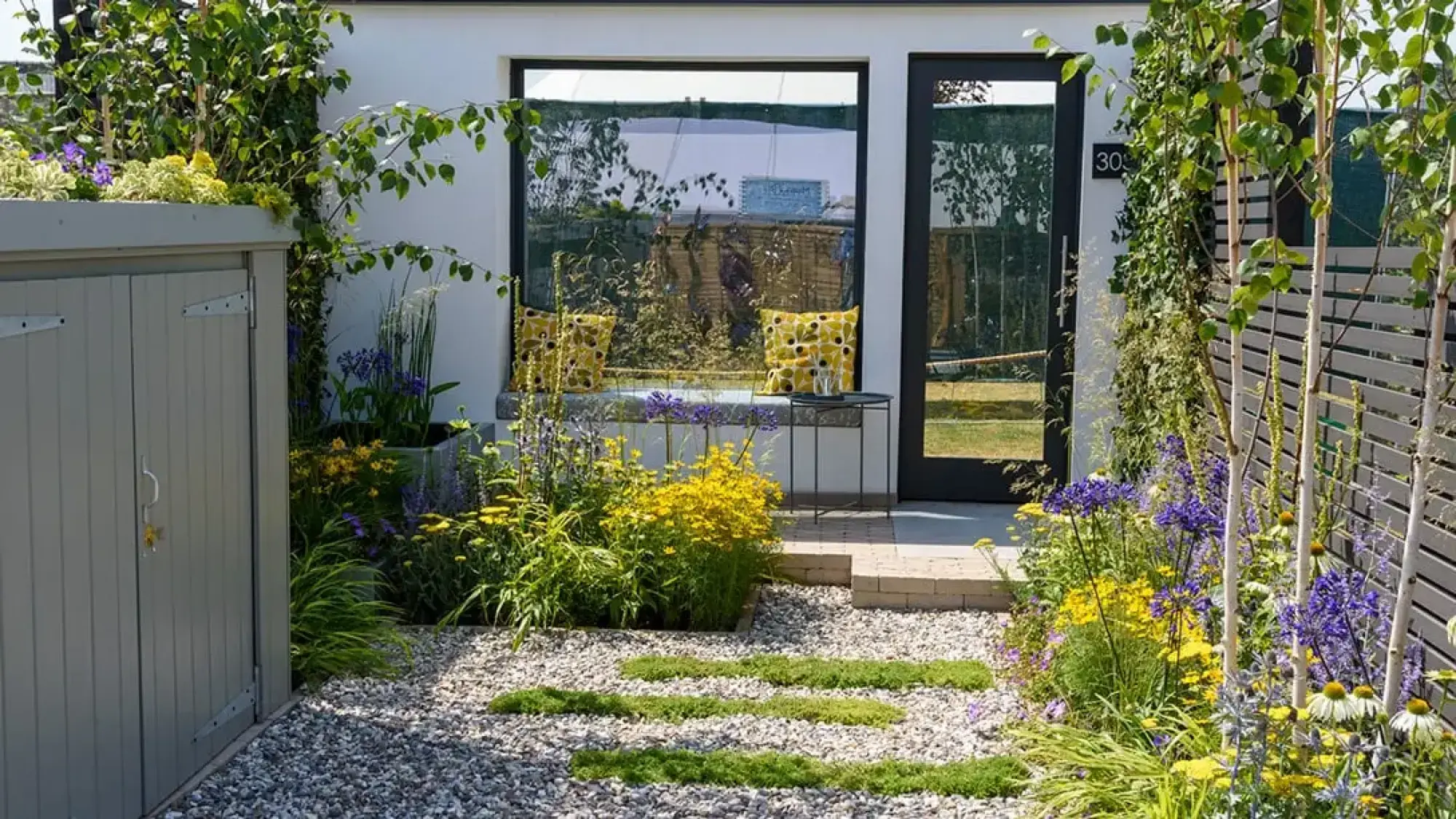 A front garden has a gravel path with moss steps leading to a window seat, framed by purple, yellow, and white flowers and a grey shed.