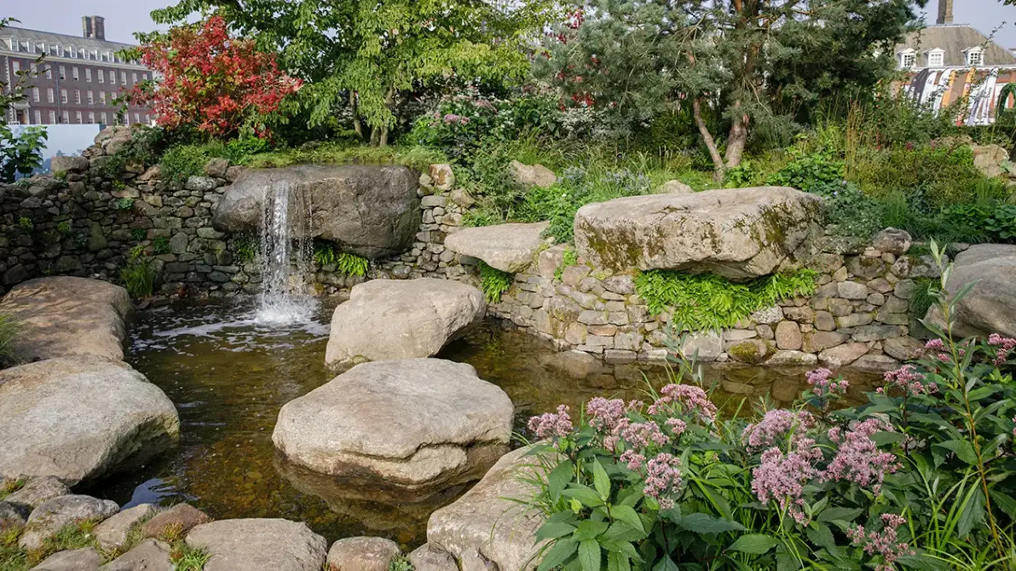 A tranquil sanctuary garden features a waterfall cascading over boulders into a pond, bordered by dry stone walls, ferns, and pink flowering plants.