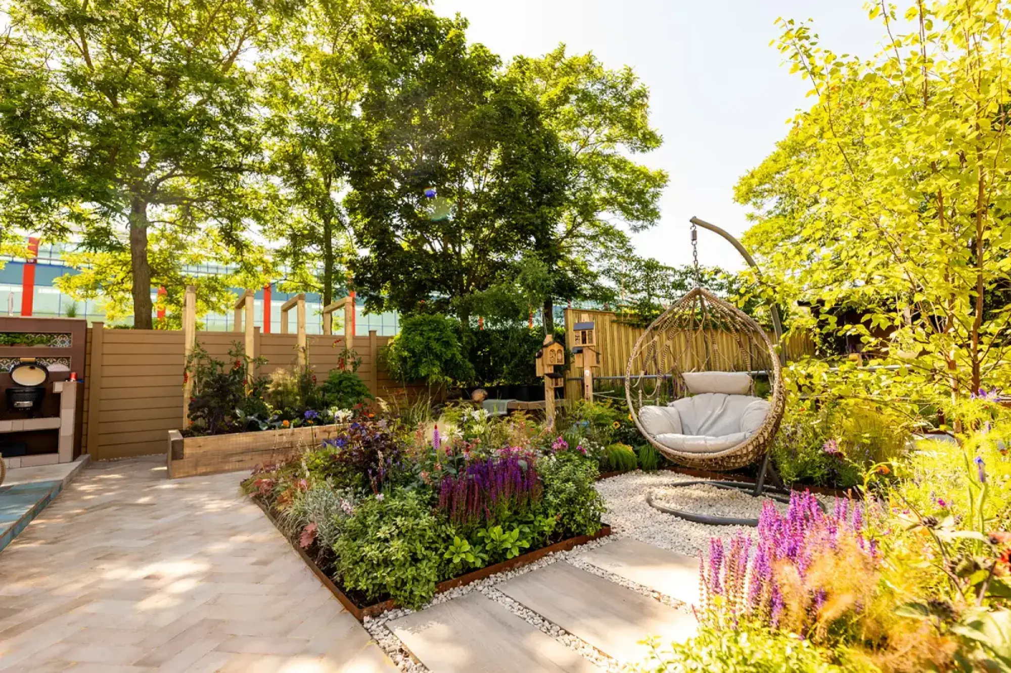 A chic show garden with a woven egg chair on gravel, a herringbone patio, an outdoor kitchen, and vibrant purple and green planting in a raised bed.