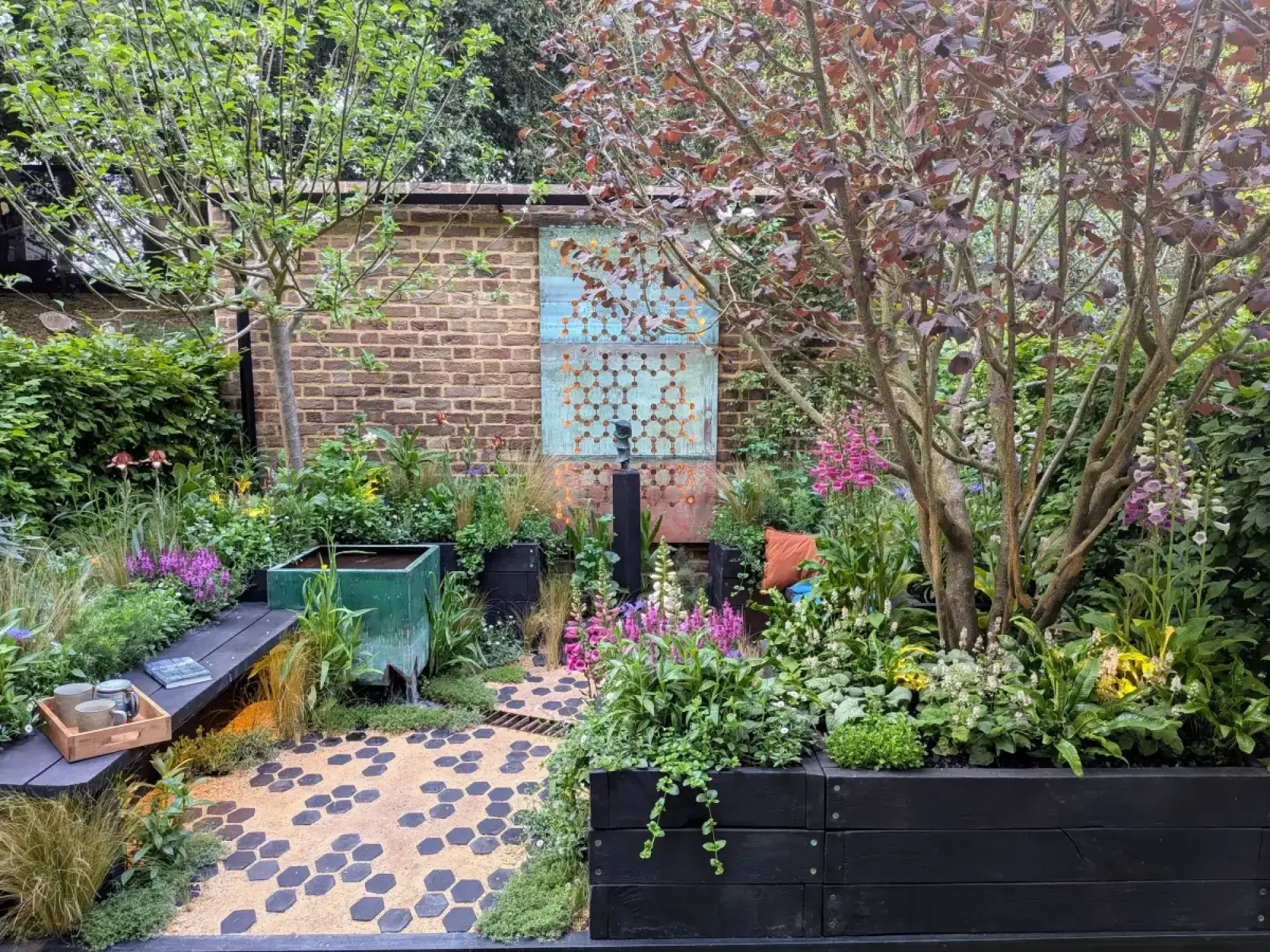 A modern container garden features verdigris copper wall art, a green water feature, black planters, a bench, hexagonal paving, and lush foxgloves.