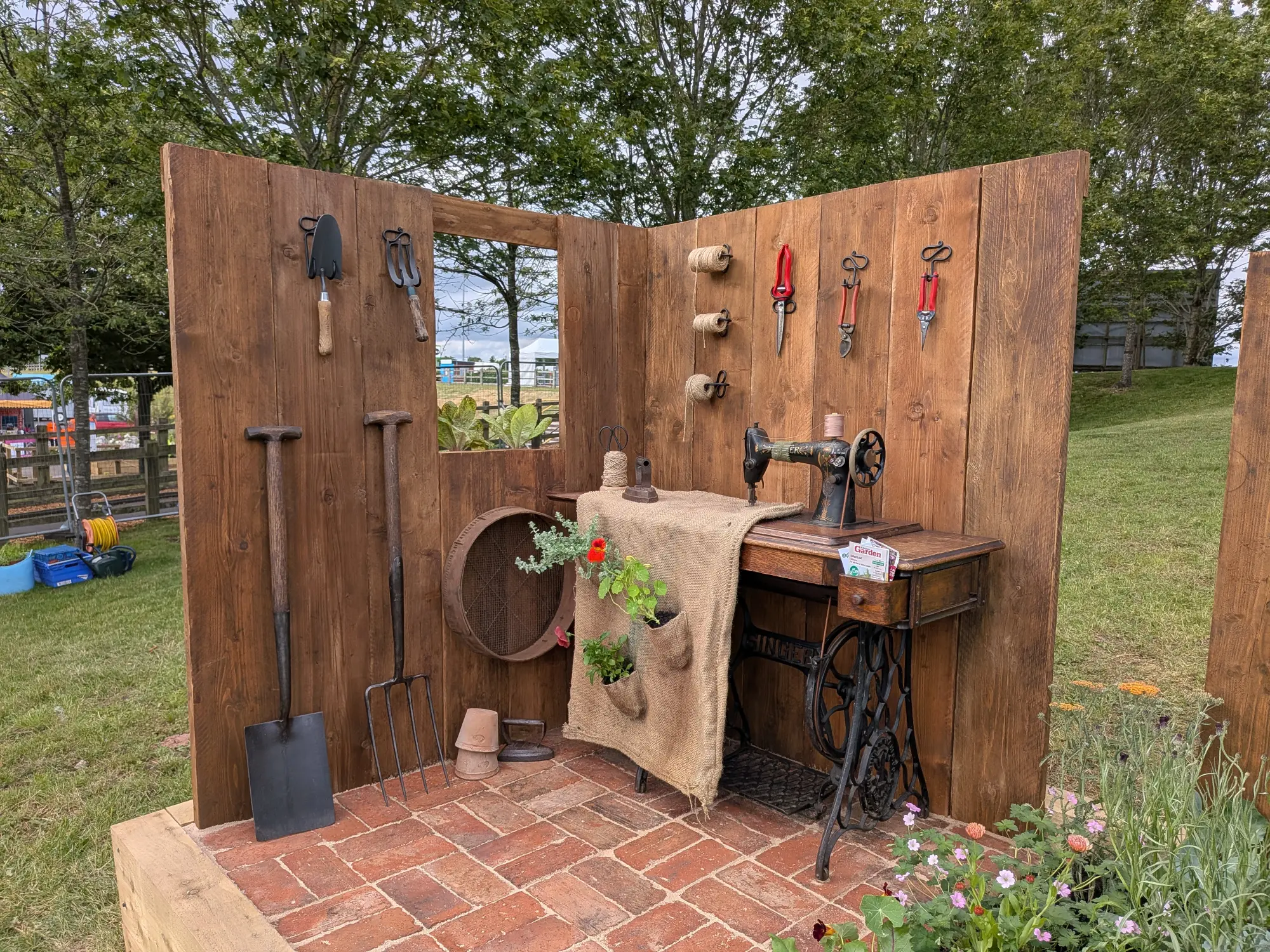 A rustic border featuring a vintage Singer sewing machine, a burlap hanging planter with flowers, and garden tools on a wooden wall and brick paved floor.
