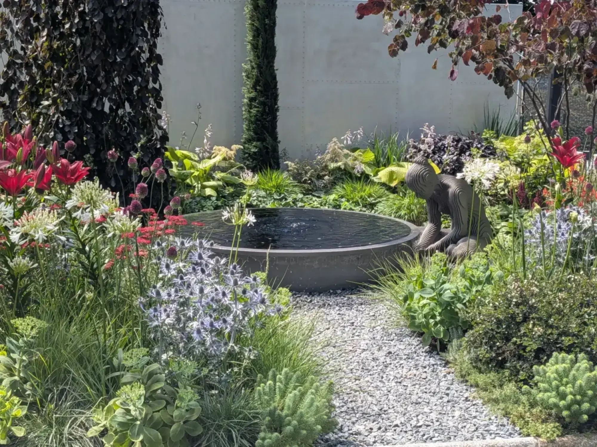 A contemporary garden featuring a dark circular water feature, a crouching layered sculpture, and a gravel path, surrounded by lush planting of red lilies, white and blue spiky flowers, and dark-leafed trees.