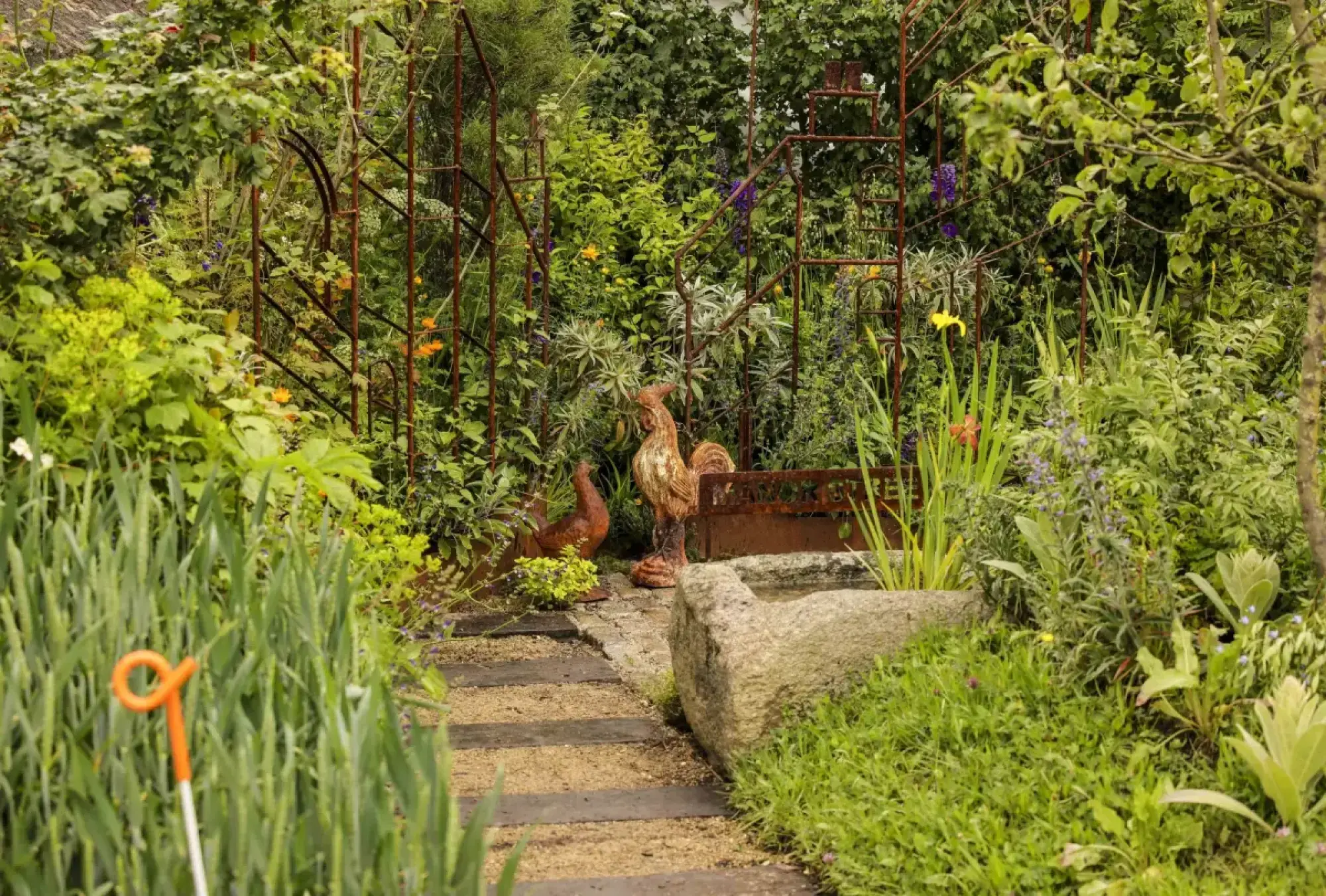 A rustic small garden features a gravel path, stone trough, and metal rooster and hen sculptures, amidst lush green plants and colourful flowers.