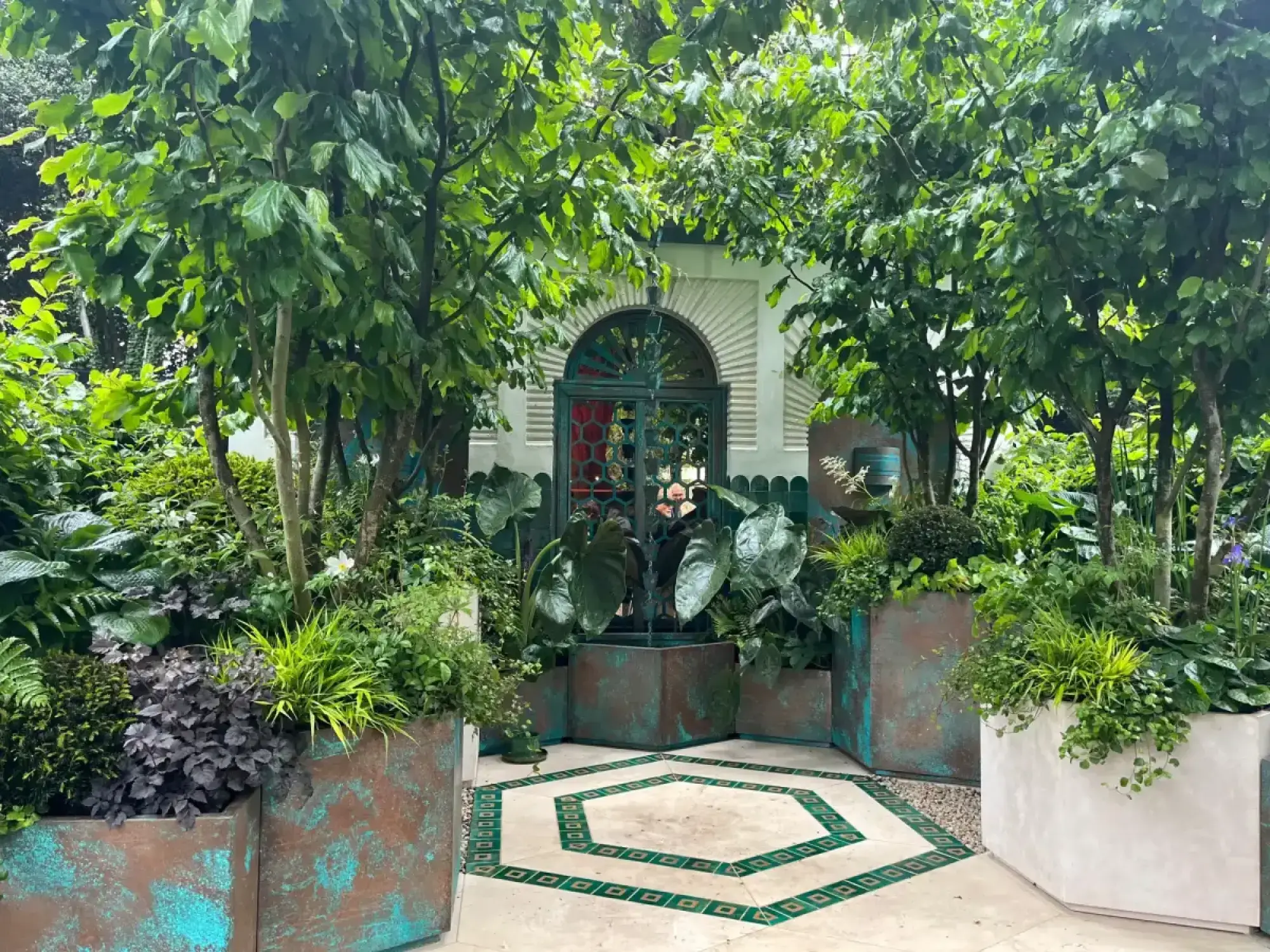 A lush container garden featuring an ornate green archway, weathered copper planters overflowing with dense green foliage, and a cream tiled patio with a green octagonal pattern.