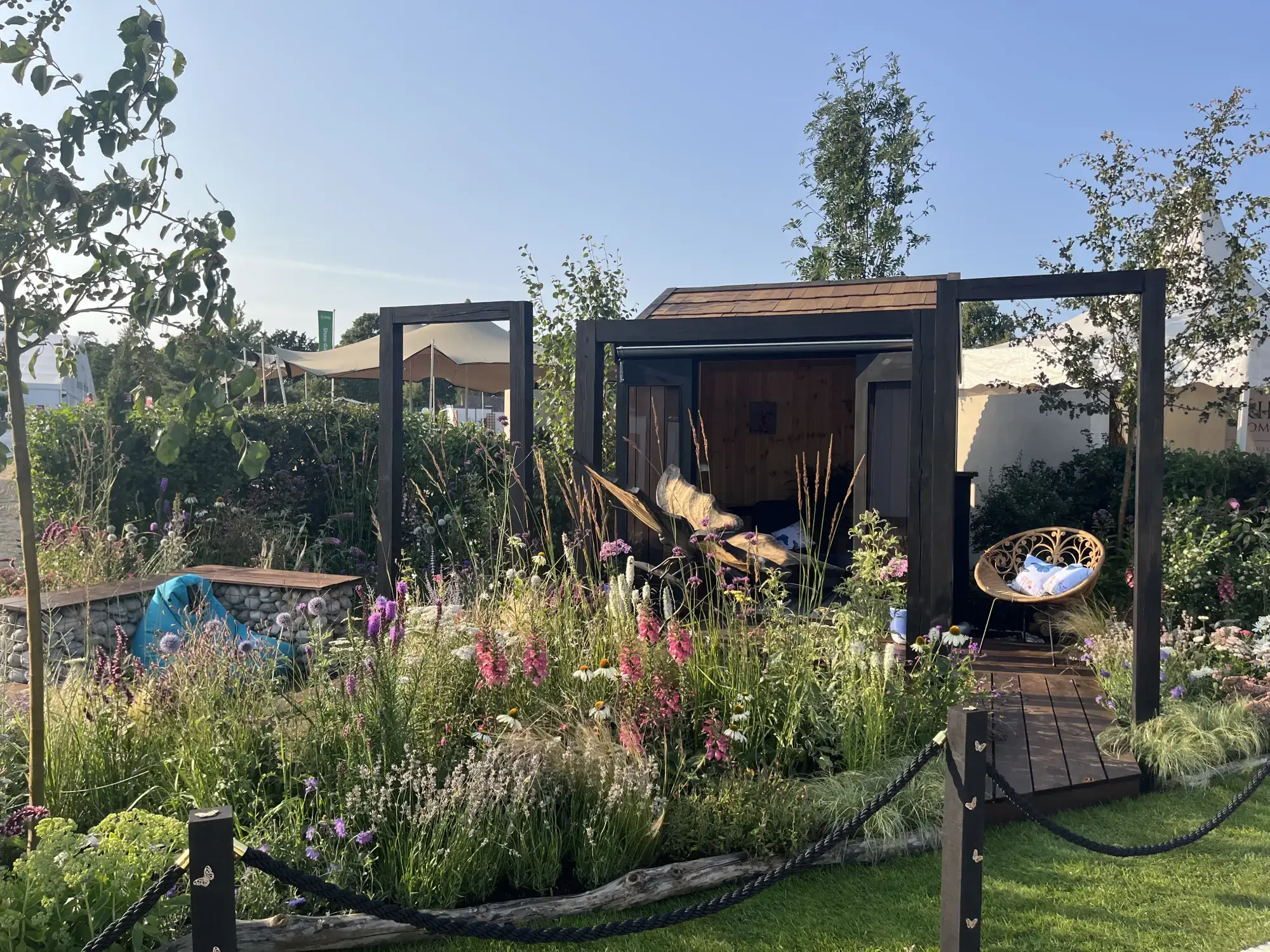 A Grow Forward Garden features a timber pod, curved lounger, and wicker chair, surrounded by pink and purple pollinator flowers.