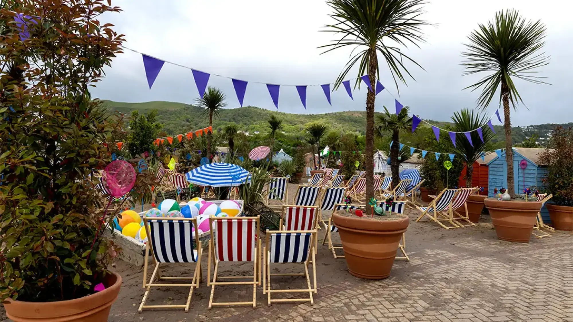 A vibrant, beach-themed garden featuring striped deck chairs, beach balls, several palm trees in terracotta pots, and colourful beach huts on sand, with bunting strung overhead.