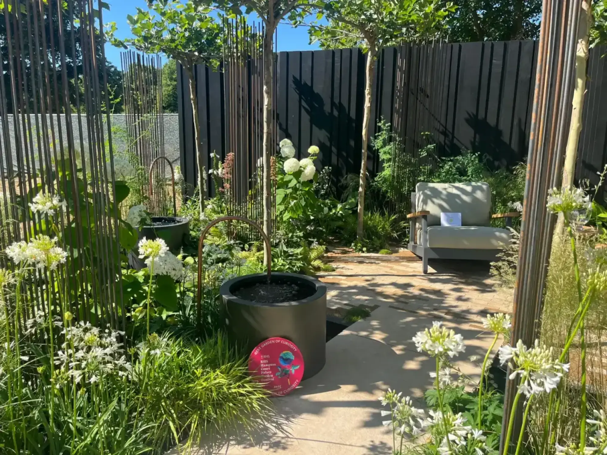 A contemporary garden features a copper-arch water feature, tall metal screens, a light grey armchair, and white agapanthus and hydrangeas on a paved path.