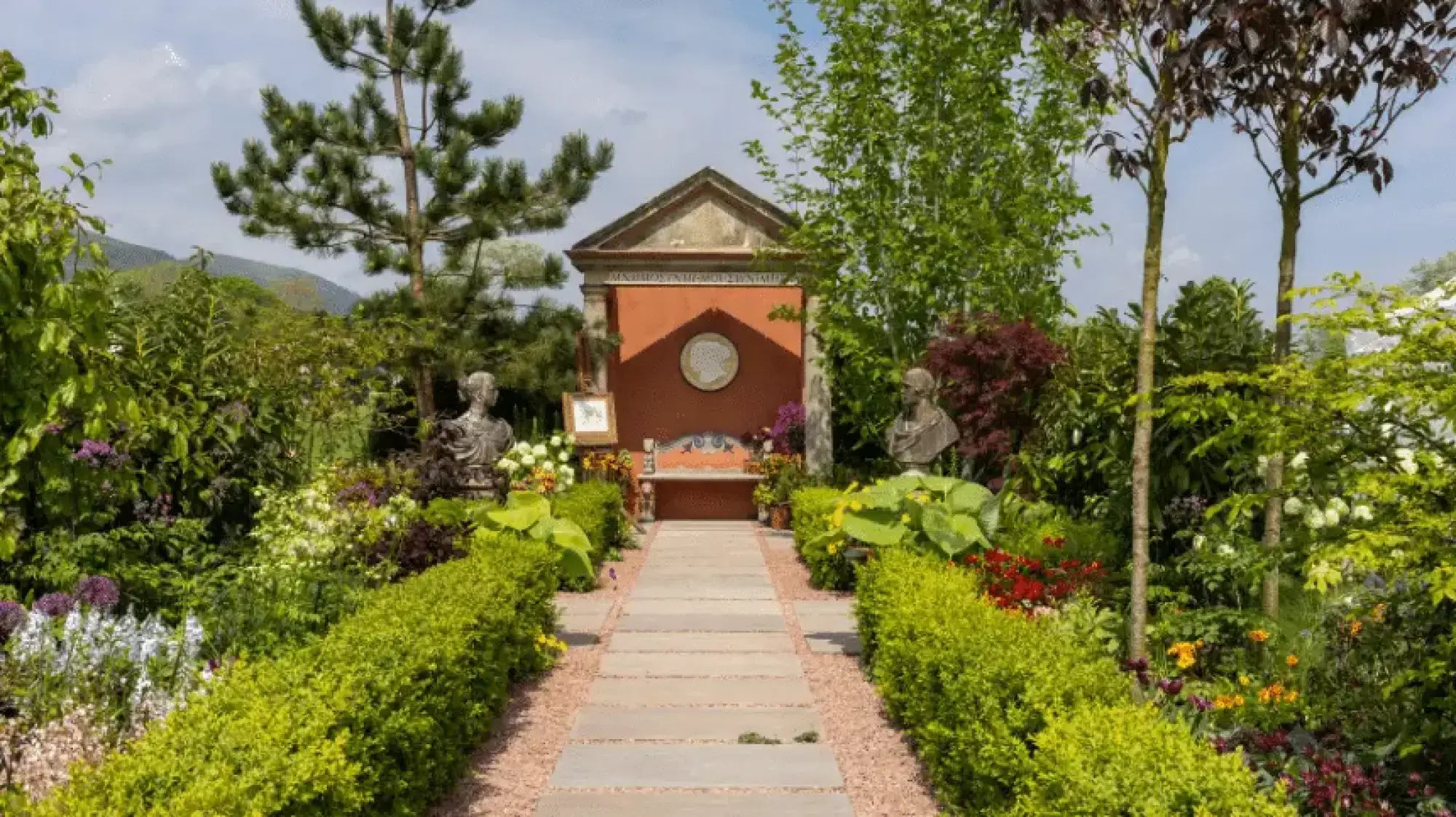 A formal classical-style garden features a paved path leading to a terracotta-red temple-style folly with an ornate bench and flanked by classical busts, set amidst lush green hedges and vibrant mixed flower borders.