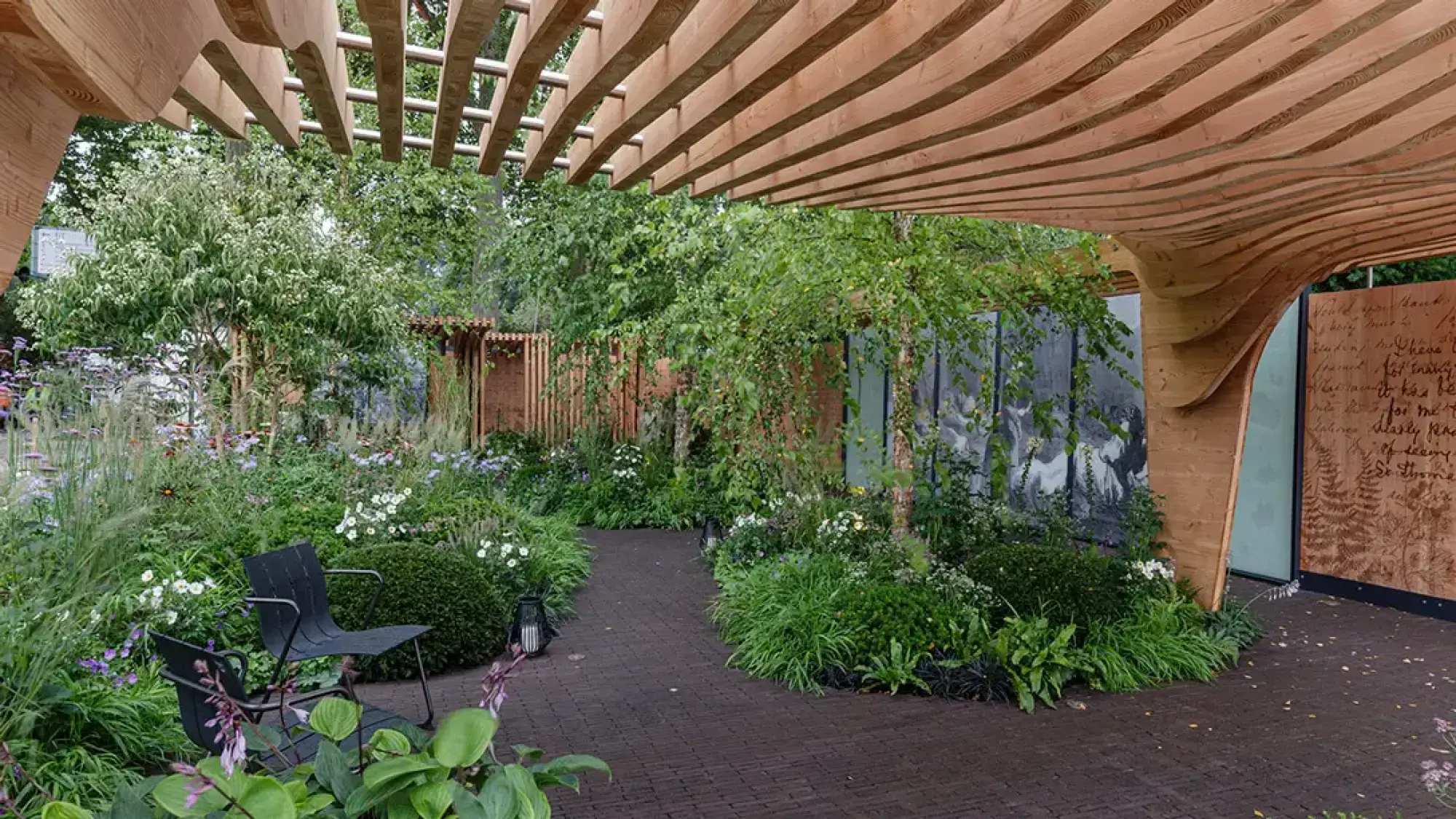 A curving timber pergola shades a winding brick path in a lush show garden with black chairs and screens of historical images.
