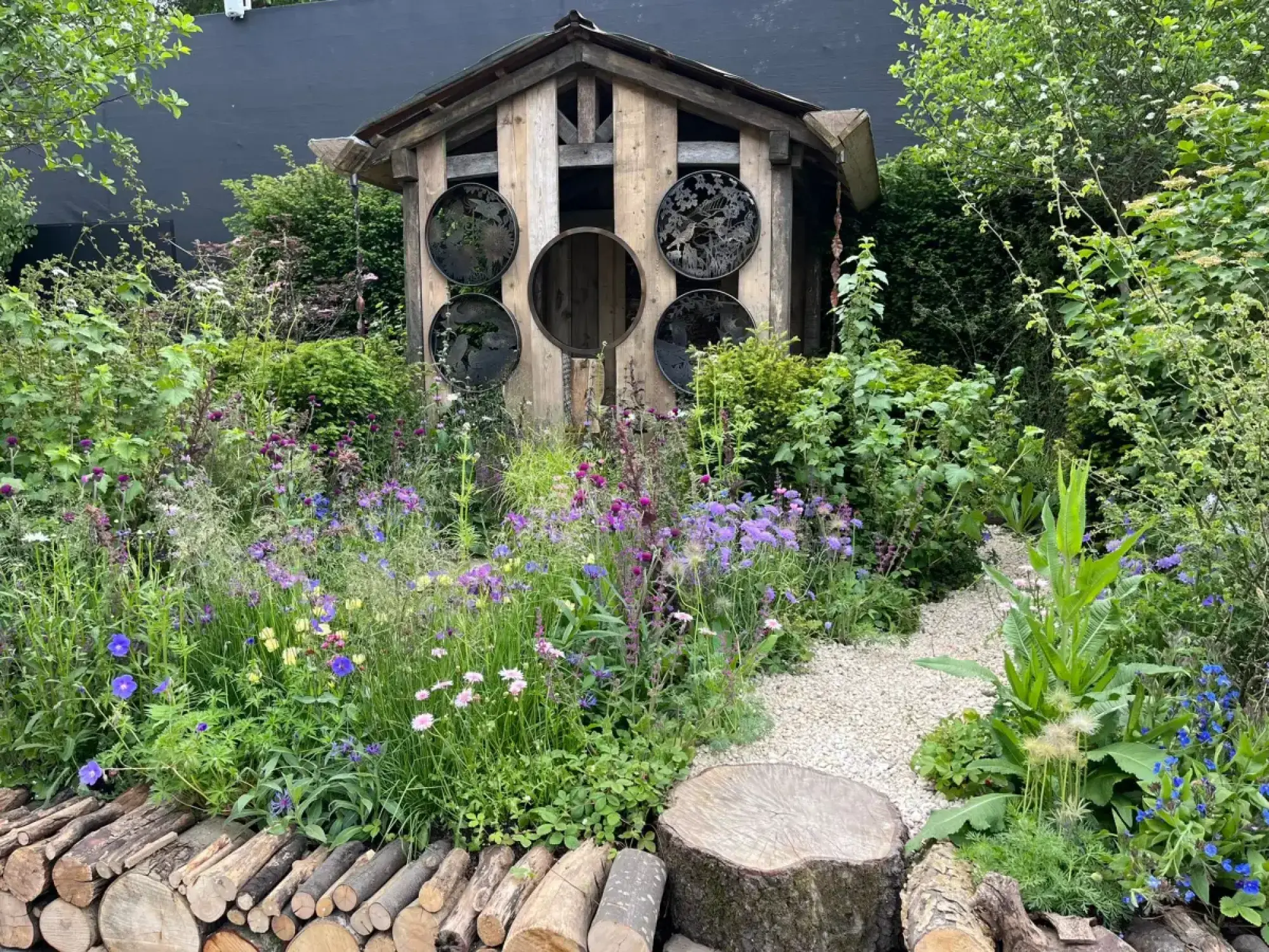 A rustic wooden bird shelter with circular metal art stands among a vibrant wildflower meadow, bordered by a log pile and a gravel path.