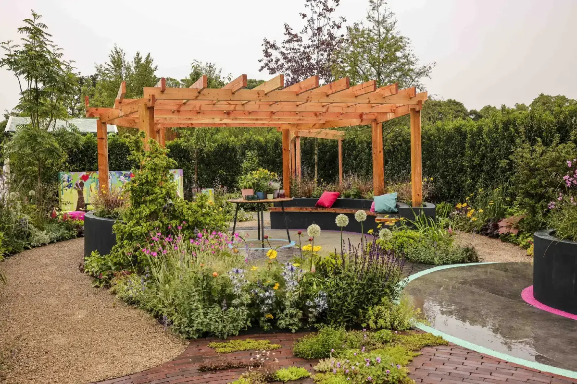 A large show garden features a timber pergola shading a black curved bench, surrounded by lush, colourful mixed planting and a gravel path leading to a circular patio with bright pink and green accent lines.