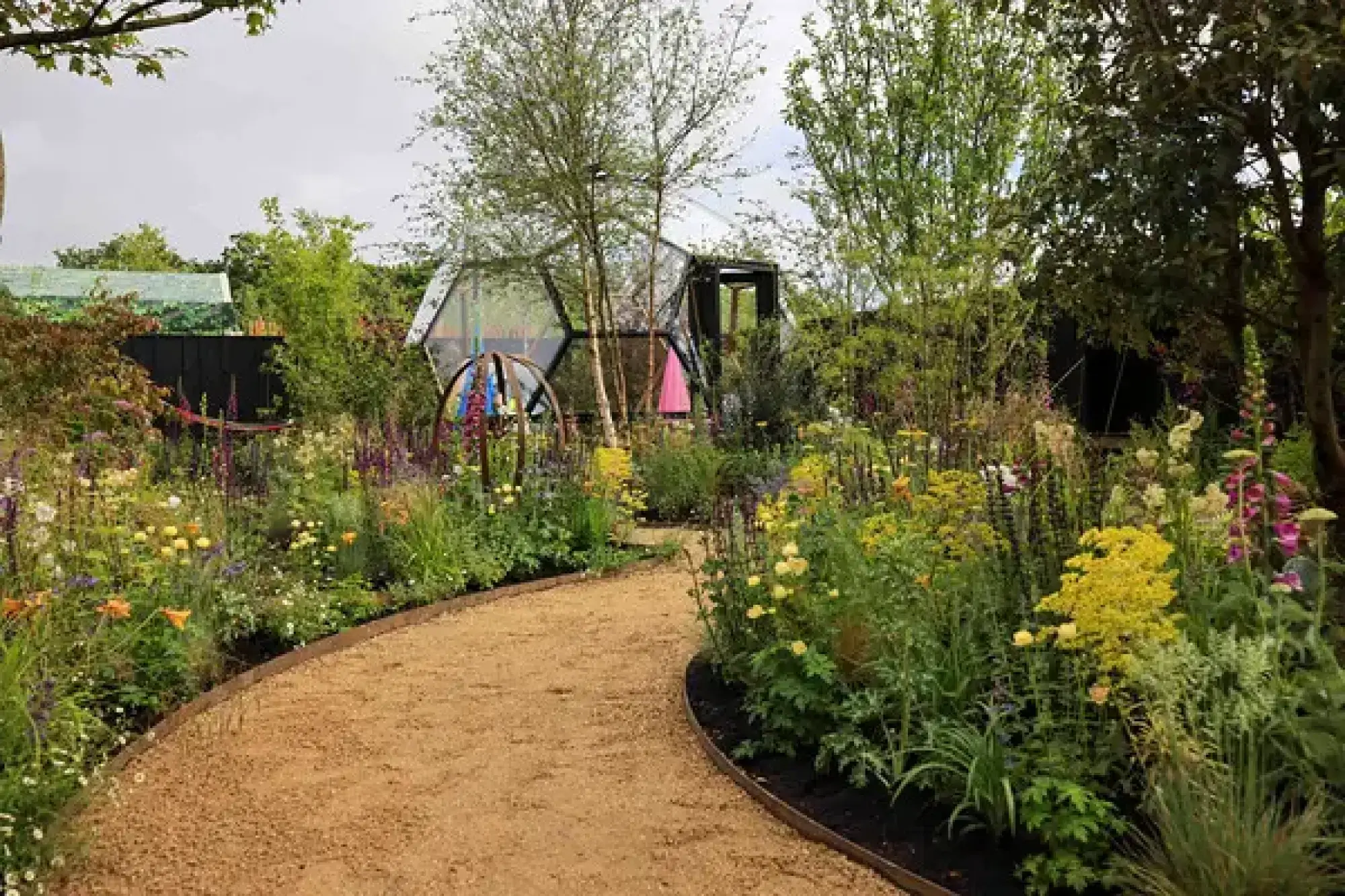 A gravel path winds through a naturalistic garden with vibrant perennials and birch trees, leading to a geodesic dome and a colourful metal sculpture.