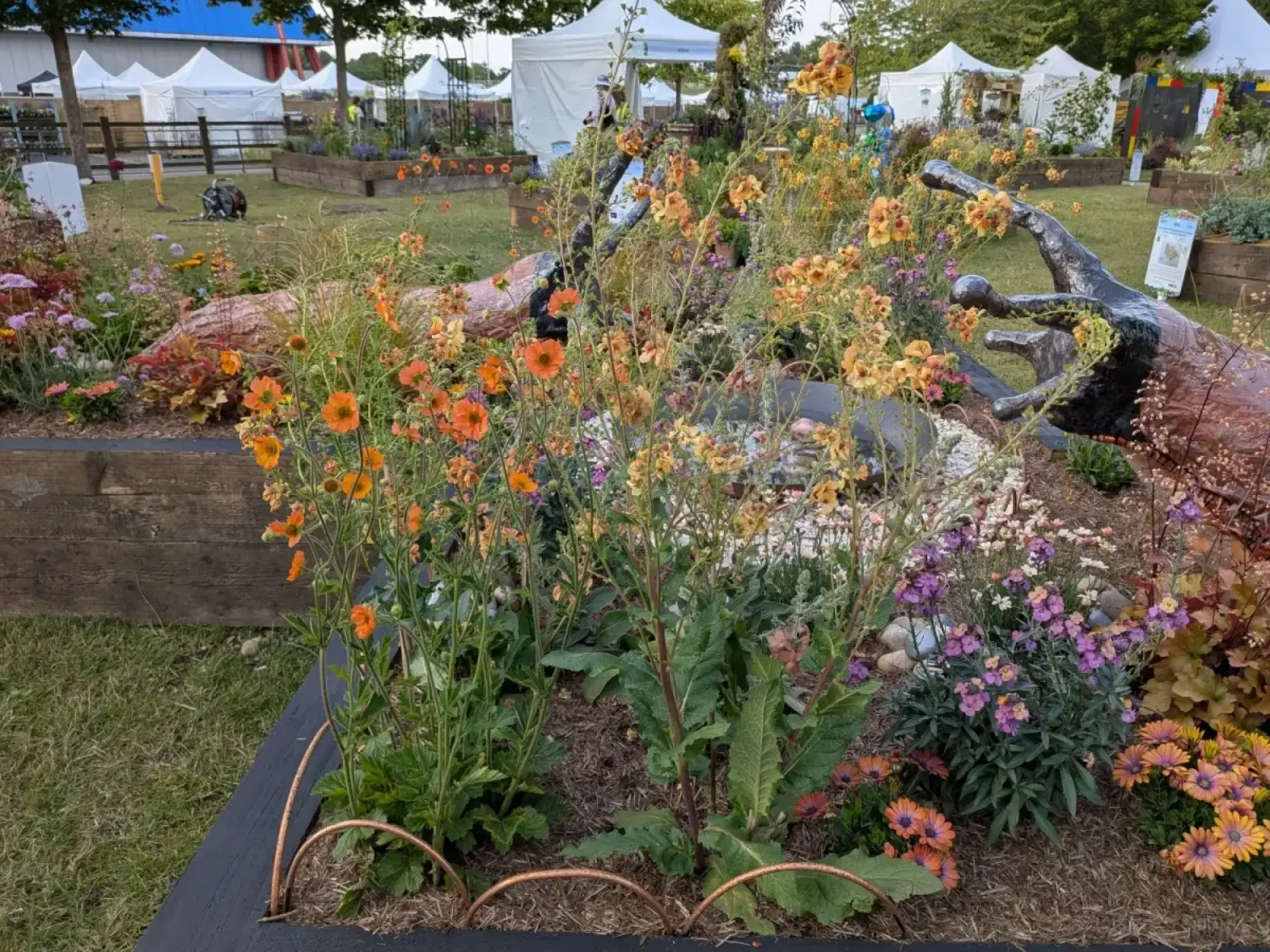 A vibrant raised garden border features a large dark slug sculpture, copper rings, and a colourful display of orange and purple flowers.