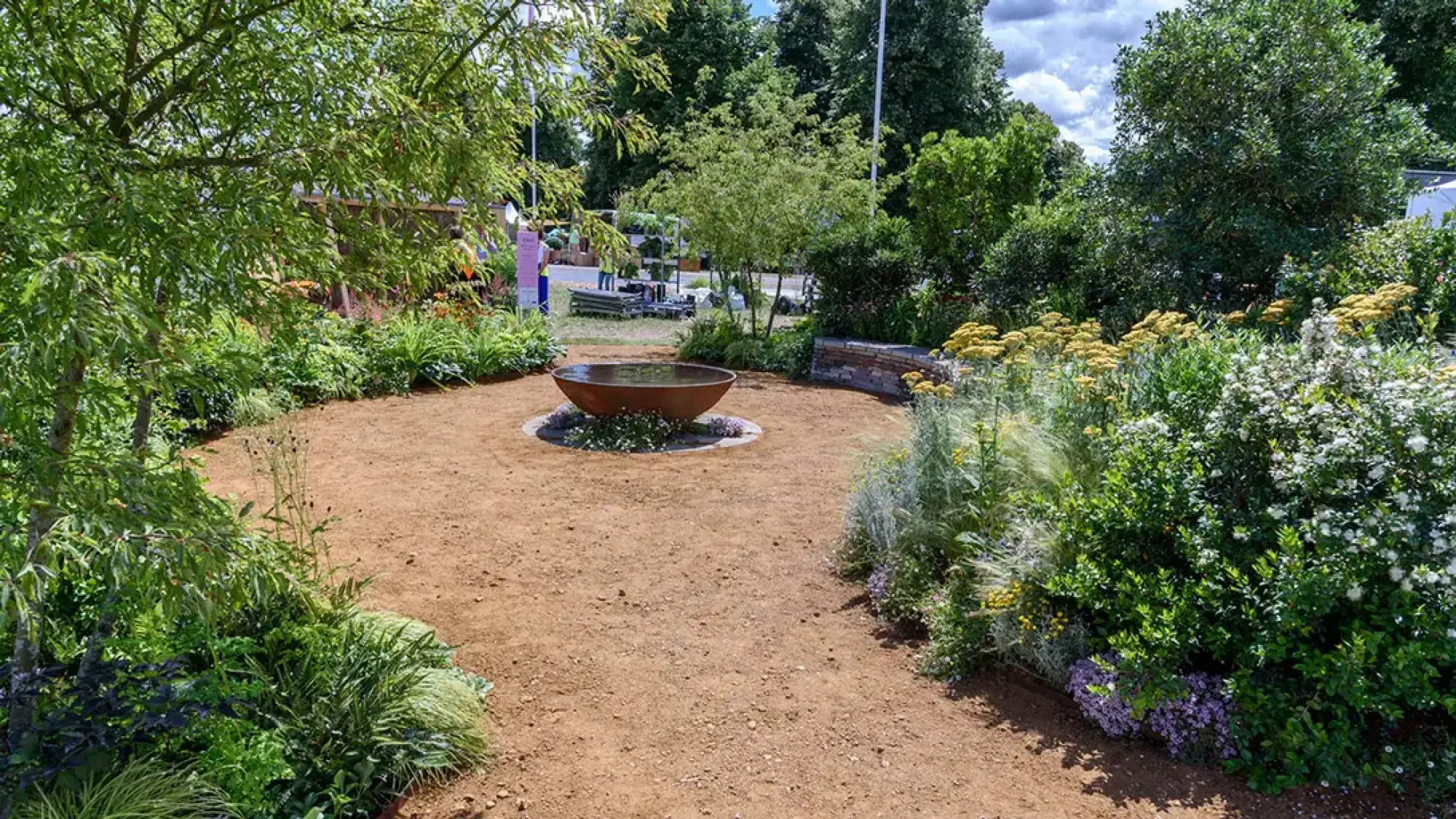 A circular garden features a large rust-coloured water bowl in the center of a bare earth area, surrounded by lush green foliage and mixed flowering plants including yellow and white blooms.