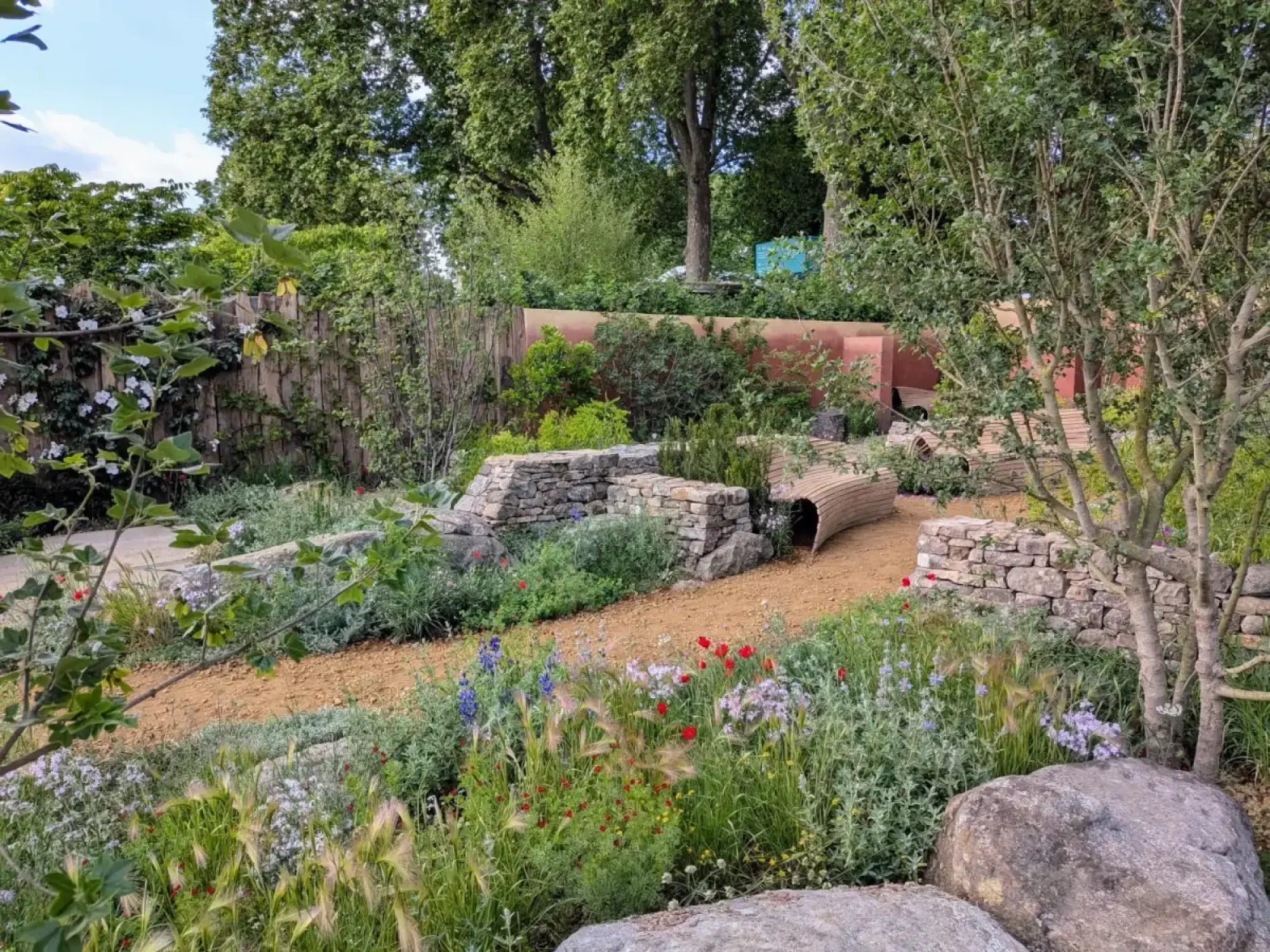 A tranquil show garden features a winding earth path, dry stone walls, a curved timber tunnel, and informal planting with red poppies, blue delphiniums, and grasses, bordered by a reddish wall.