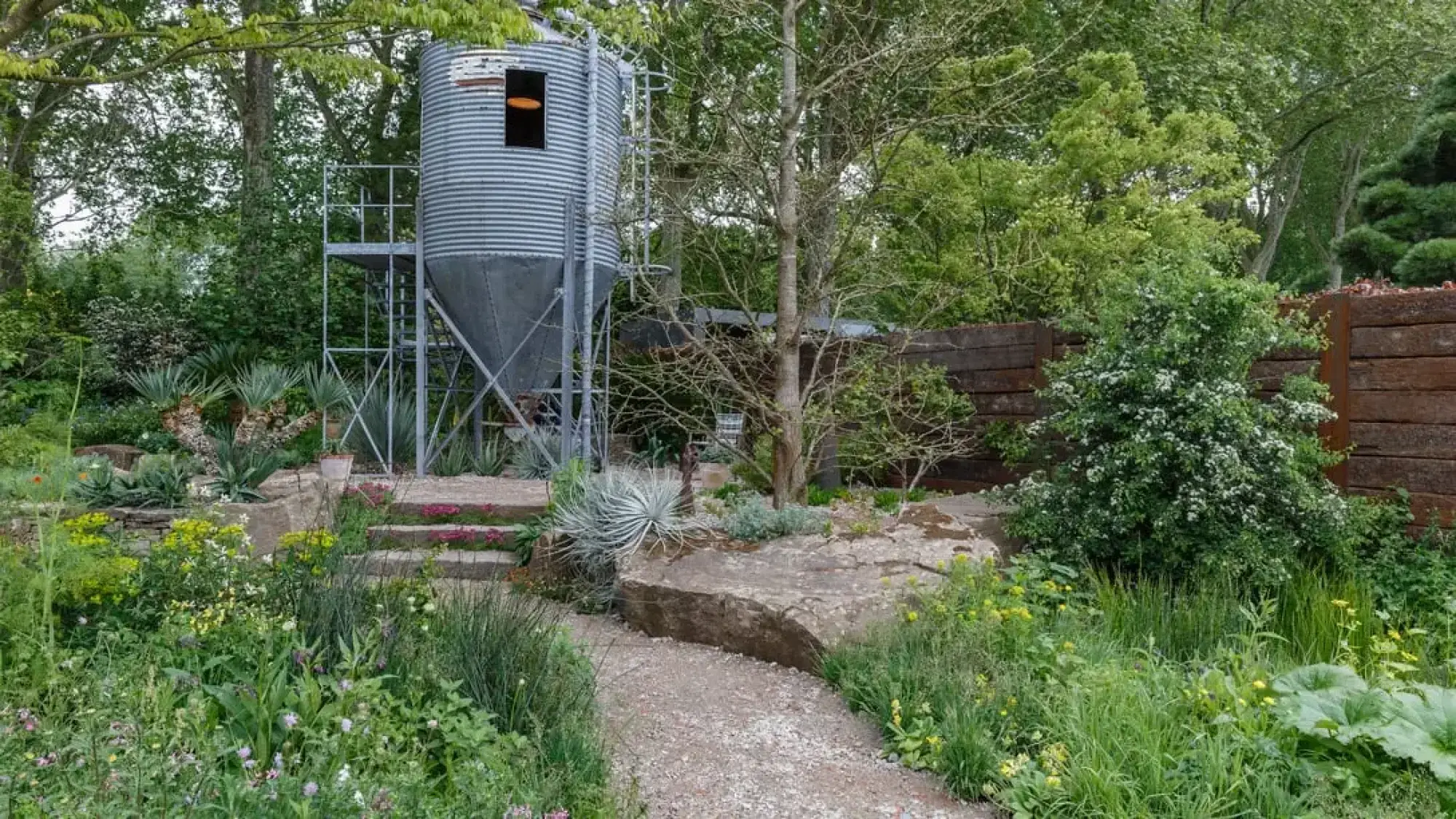 A garden features a metal grain silo, external stairs, a gravel path, boulders, and terraced planting of succulents and wildflowers.