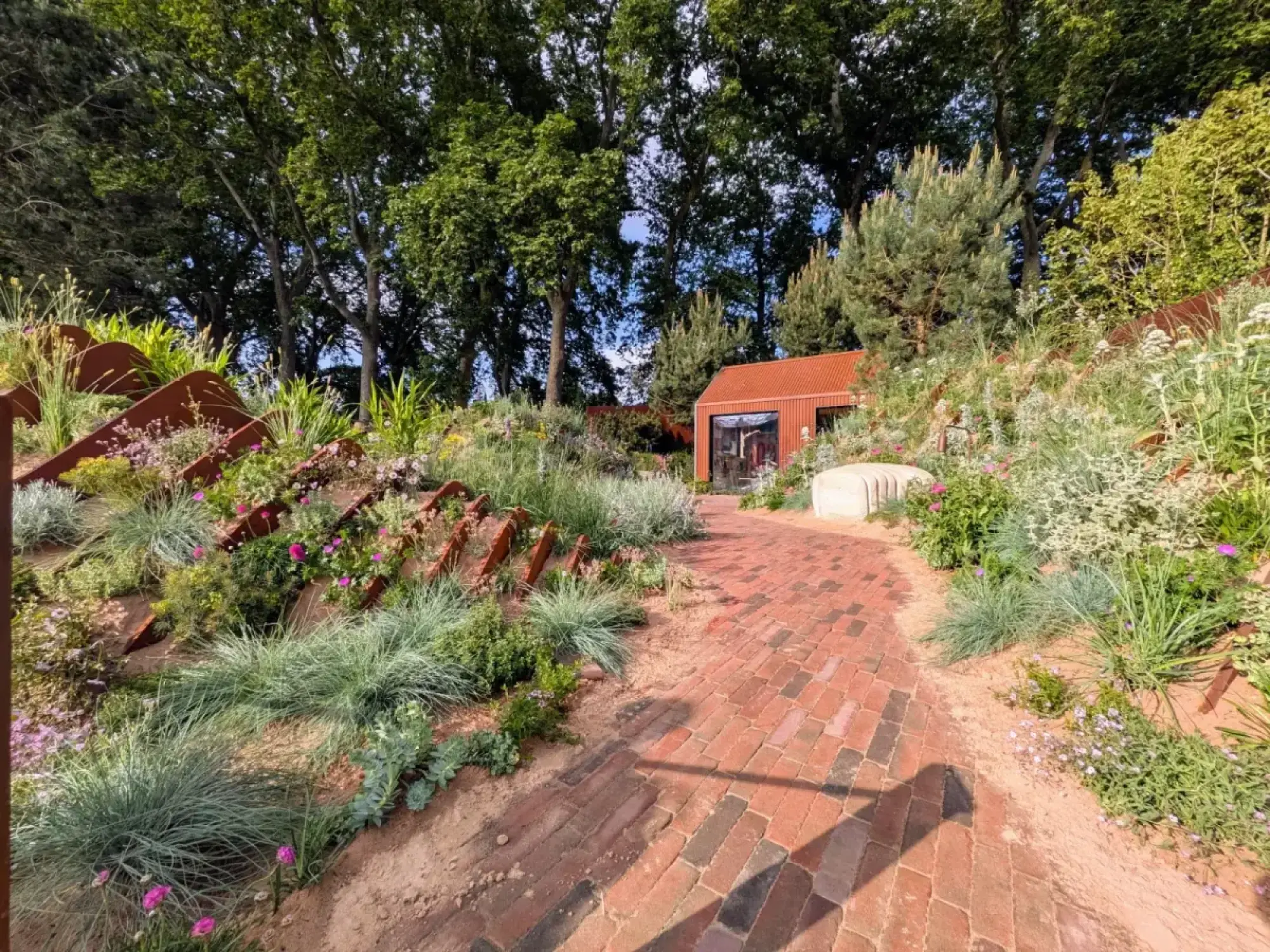A red brick path leads through a contemporary show garden featuring Corten steel retaining walls, drought-tolerant planting, and a red corrugated metal cabin in the distance.