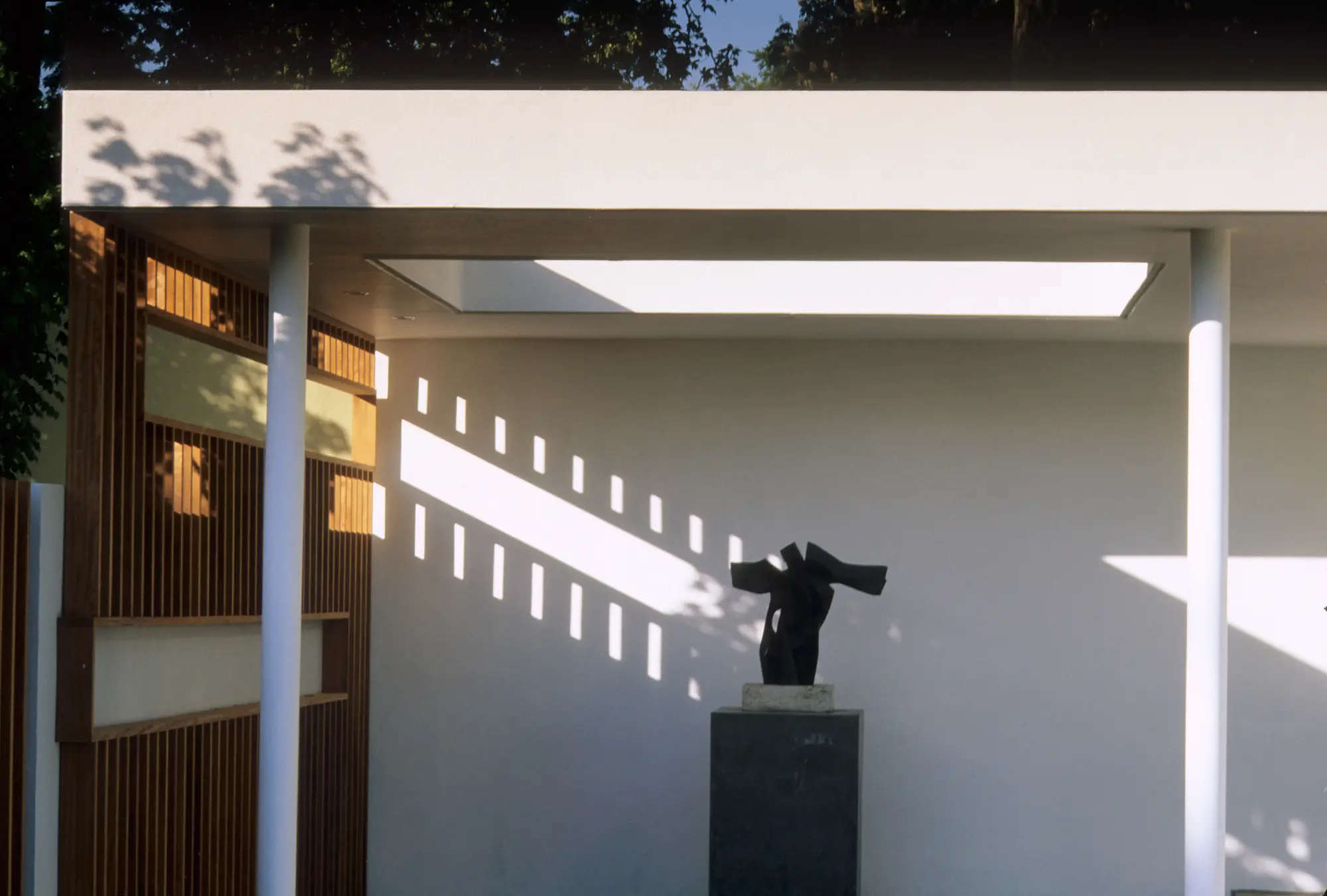 A modern show garden featuring a black abstract sculpture on a dark plinth, set against a white wall, under a white canopy supported by white columns, alongside a wooden slatted screen with recessed shelving.