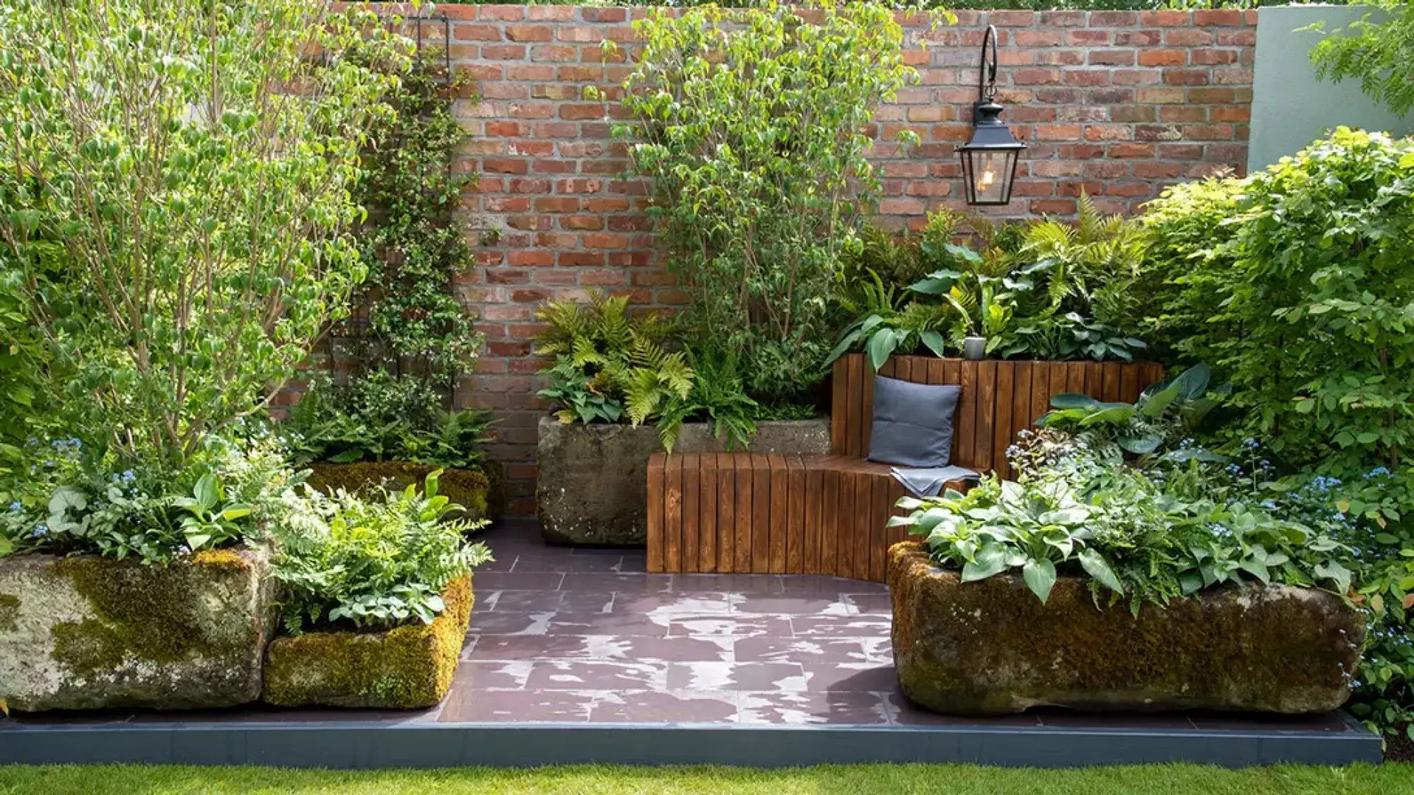 A tranquil container garden features a slatted timber bench against a brick wall, flanked by lush green ferns and hostas in moss-covered stone troughs.