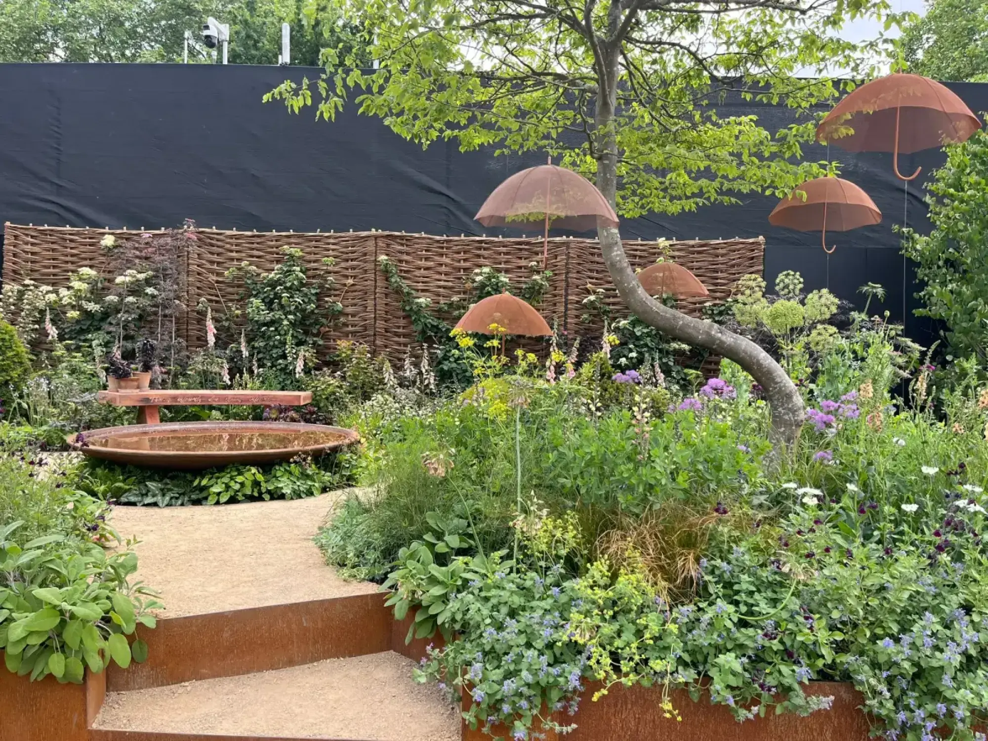 A naturalistic show garden features a gravel path, Corten steel steps, and a circular water feature, surrounded by lush mixed planting, with rusted metal umbrellas suspended from a tree and a woven hazel screen backdrop.